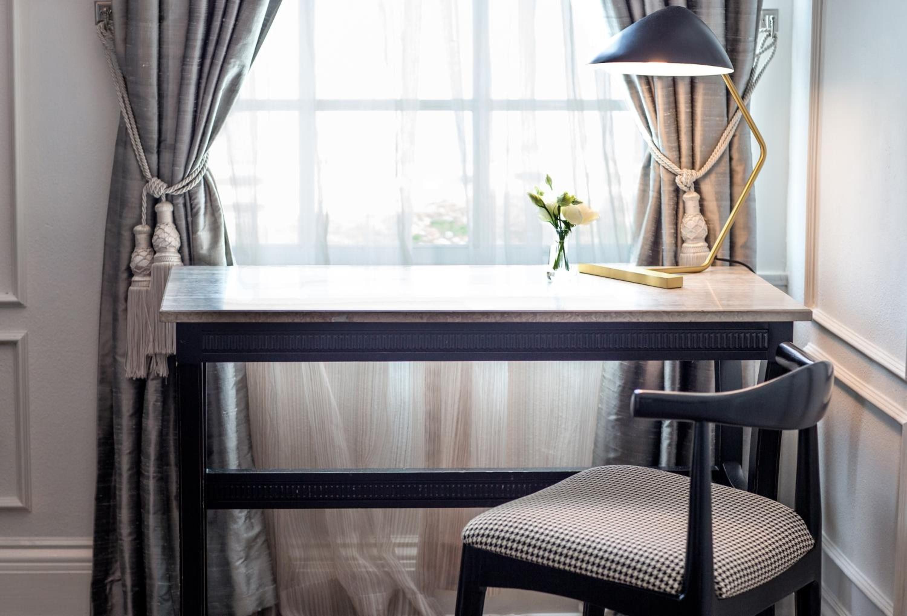 Natural light streams across a thoughtfully appointed writing desk, where brass fixtures and soft gray linens create an intimate workspace perfect for morning correspondence.