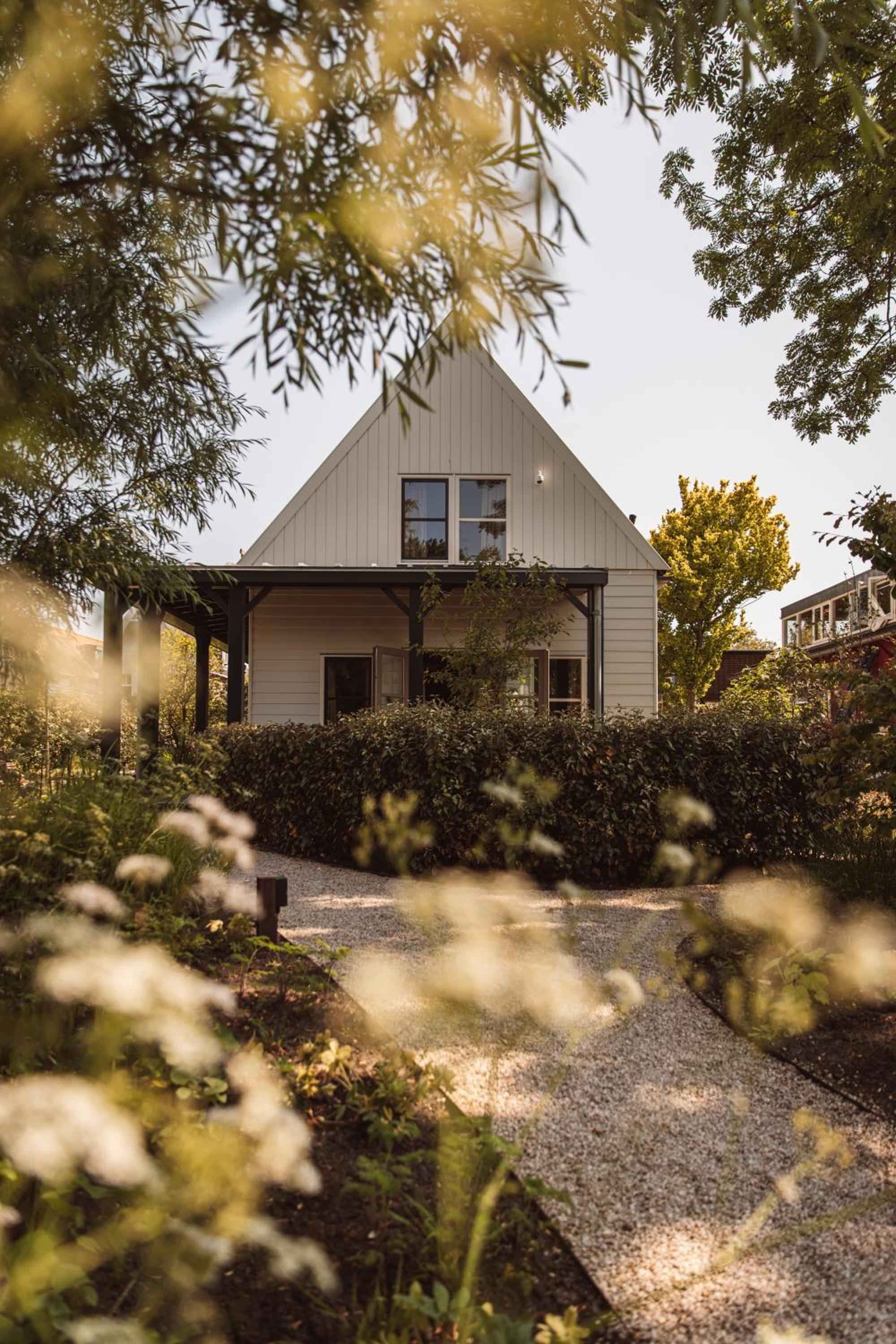 Golden hour light filters through overhanging branches, illuminating the charming white cottage's covered porch and gravel pathway lined with wild hedgerows at this intimate Dutch retreat.