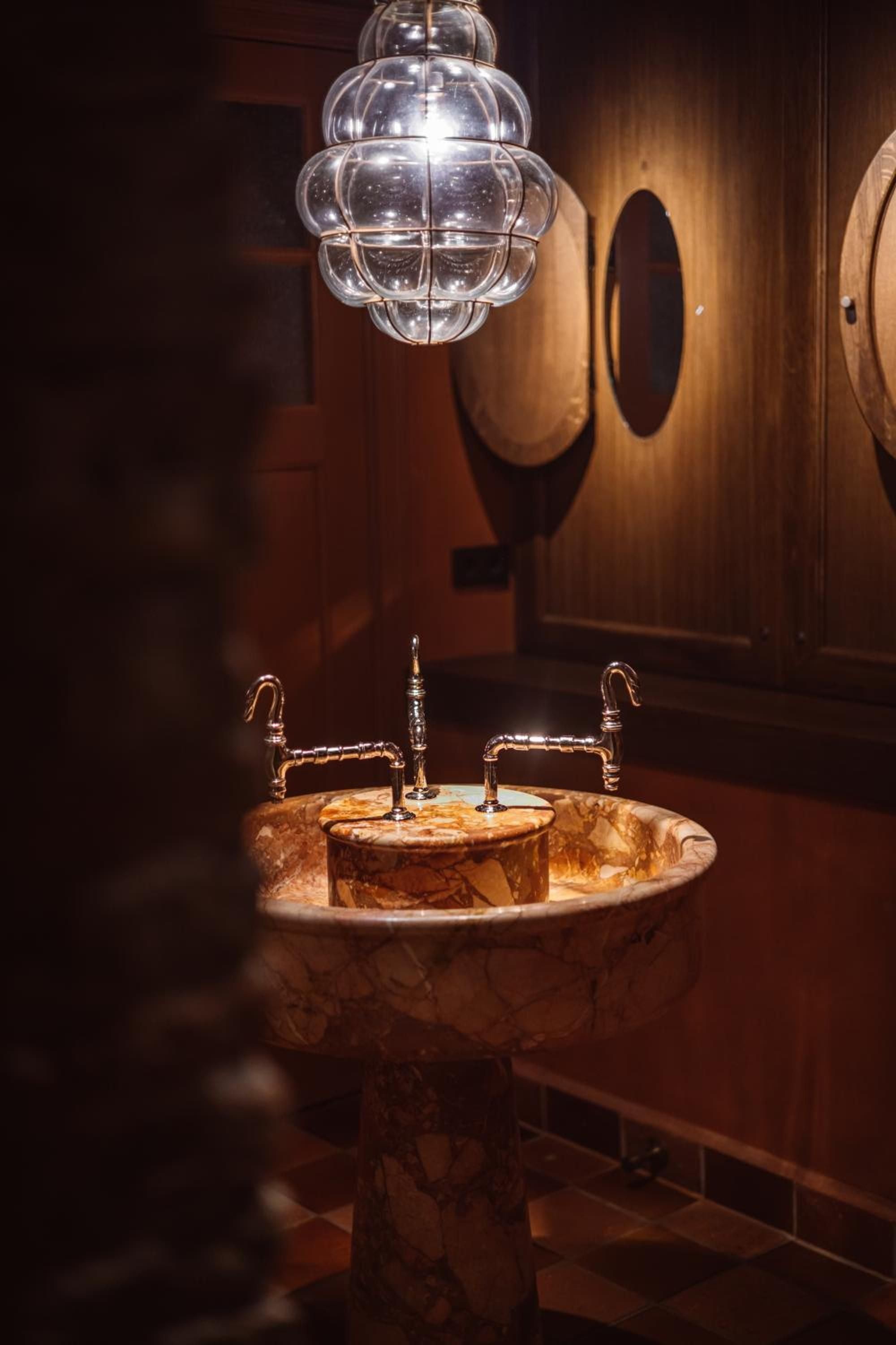 Warm amber light bathes the sculptural marble basin, where brass fixtures gleam beneath a stunning glass pendant that transforms this intimate powder room into pure artistry.