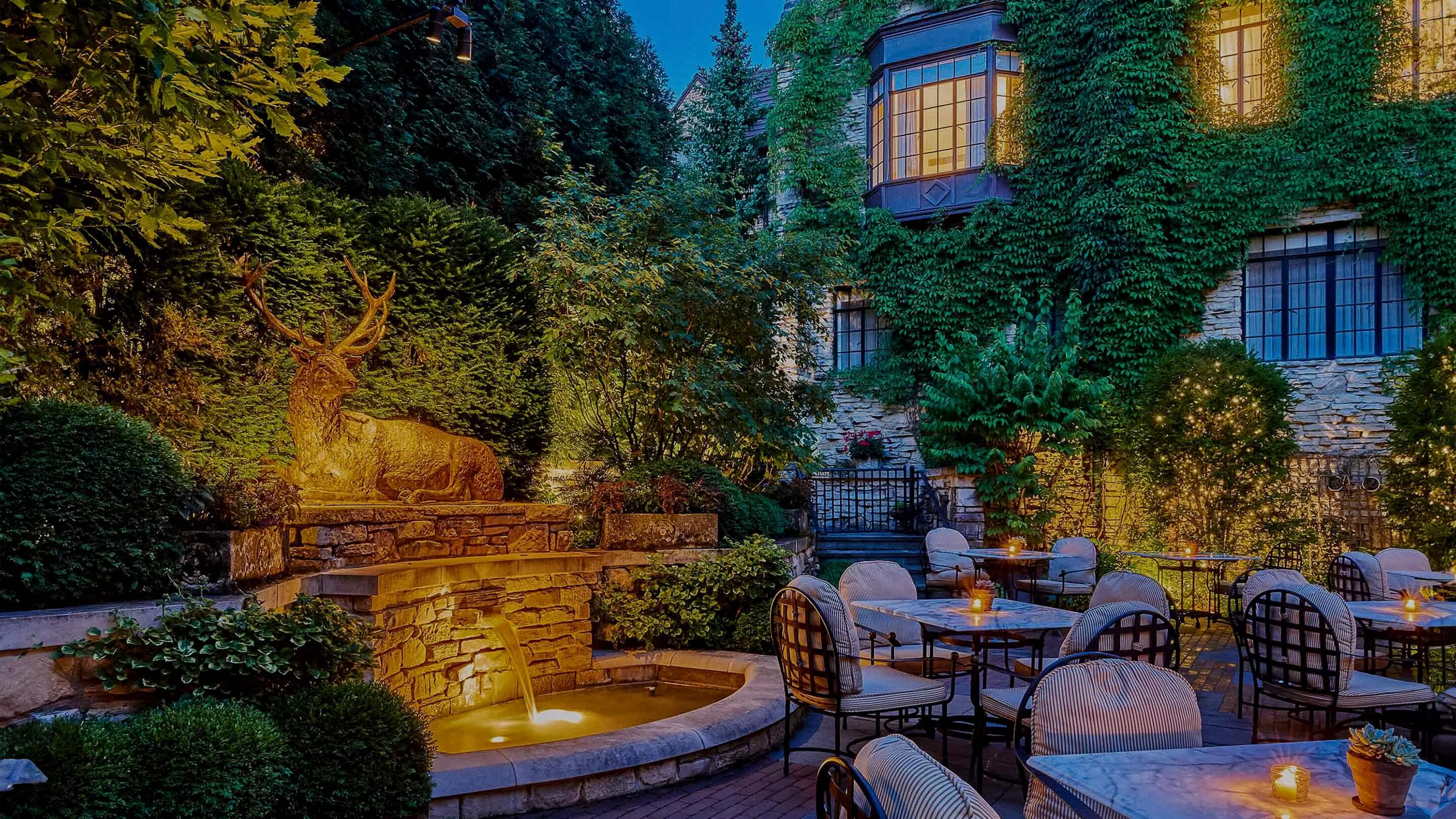 Golden hour illuminates the ivy-draped courtyard where intimate candlelit tables nestle beside a deer sculpture fountain, creating an enchanted dining refuge.