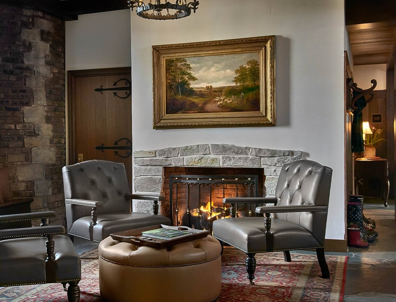 Leather wingback chairs gather around a crackling stone fireplace beneath oil paintings, while wrought-iron chandeliers cast warm light across exposed brick walls.