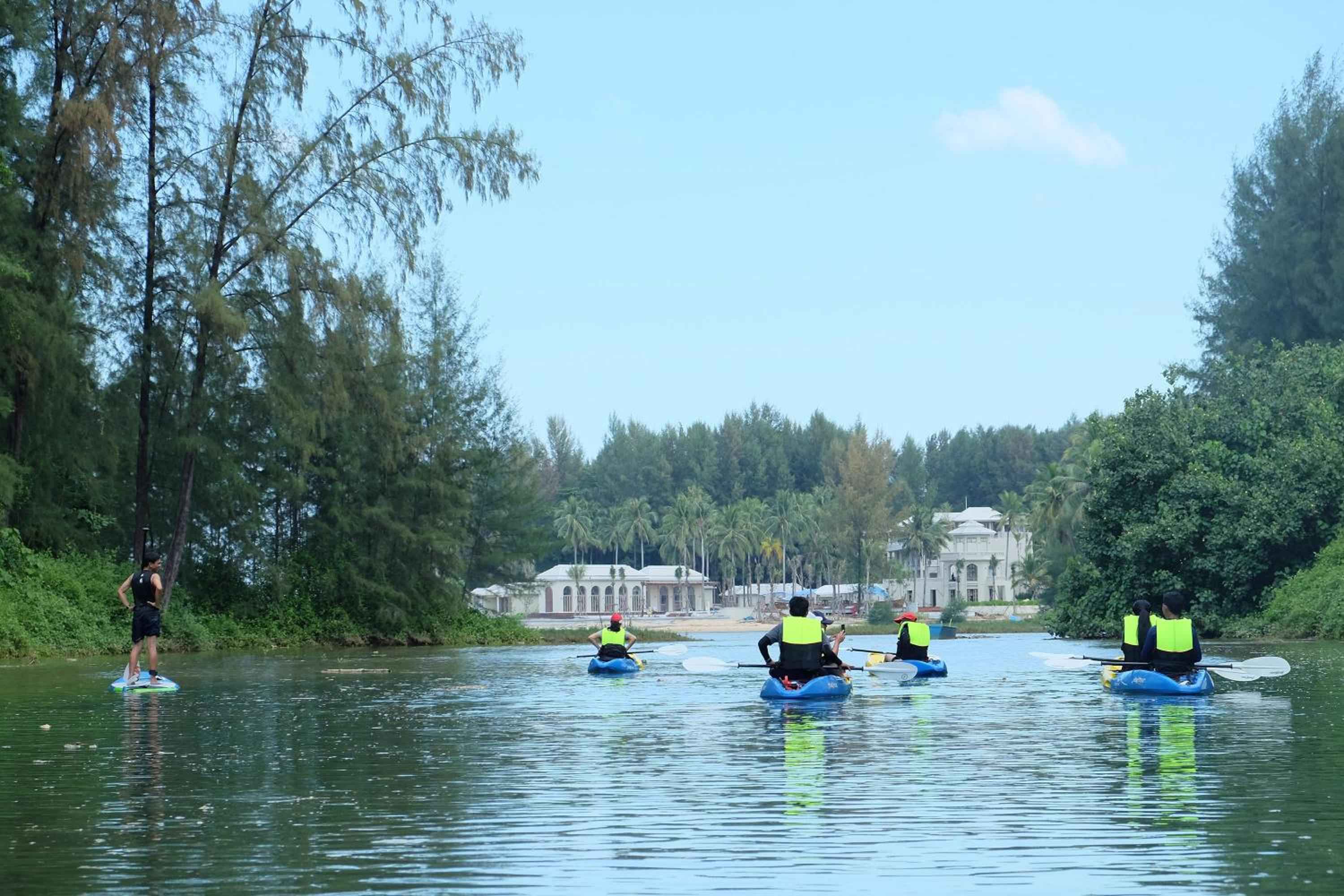 Paddlers in bright safety vests navigate tranquil waters past colonial-style architecture, while a lone stand-up paddleboarder glides beneath drooping birch branches.