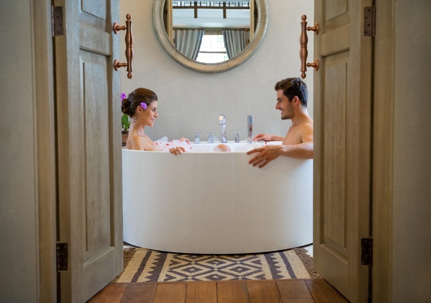 Couples unwind in the freestanding soaking tub, where rose petals float on warm water and carved wooden doors frame this intimate spa-like sanctuary.