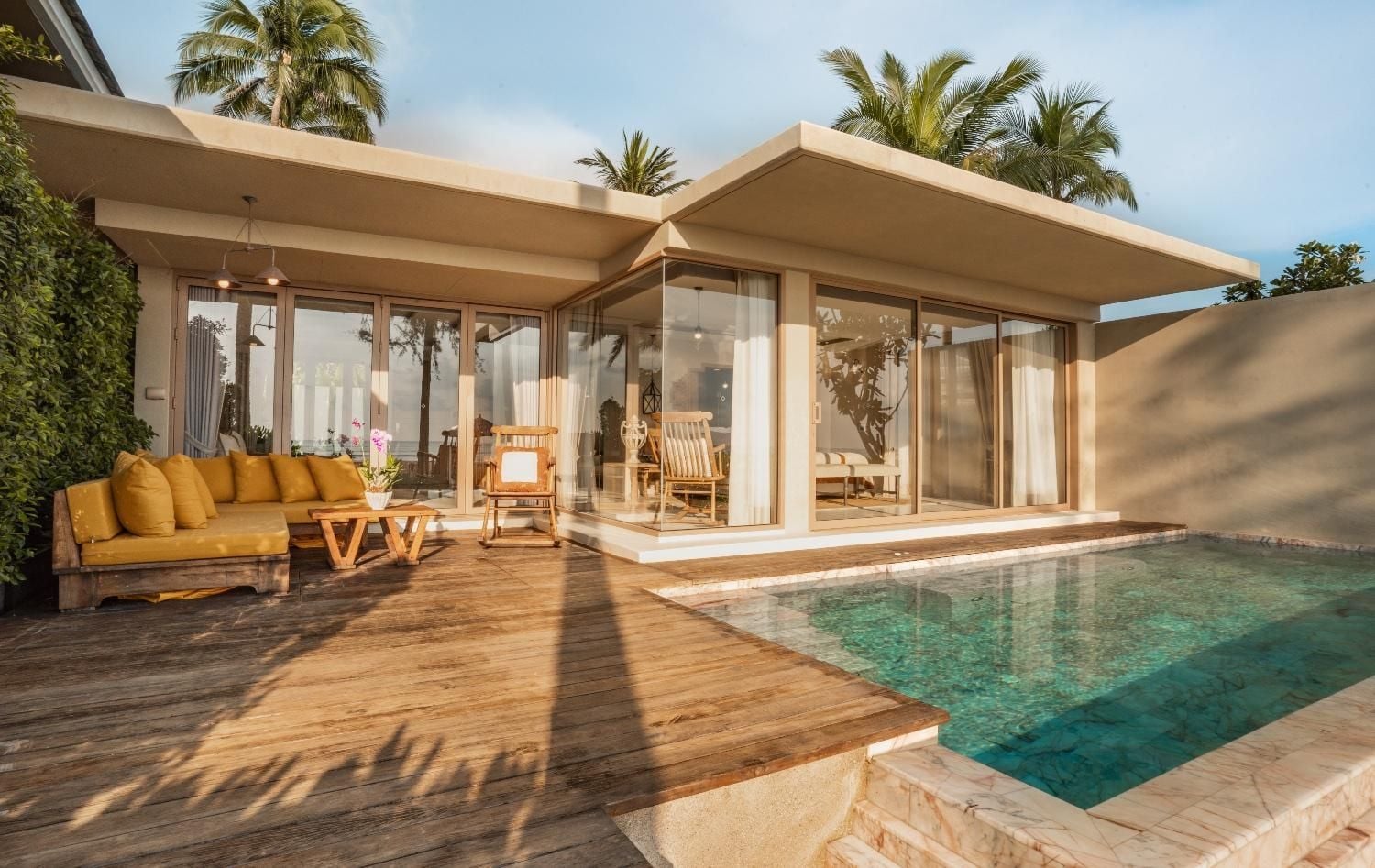 Golden hour light filters through swaying palms onto warm teak decking, where mustard cushions invite lingering beside crystalline waters that mirror the tropical sky above.