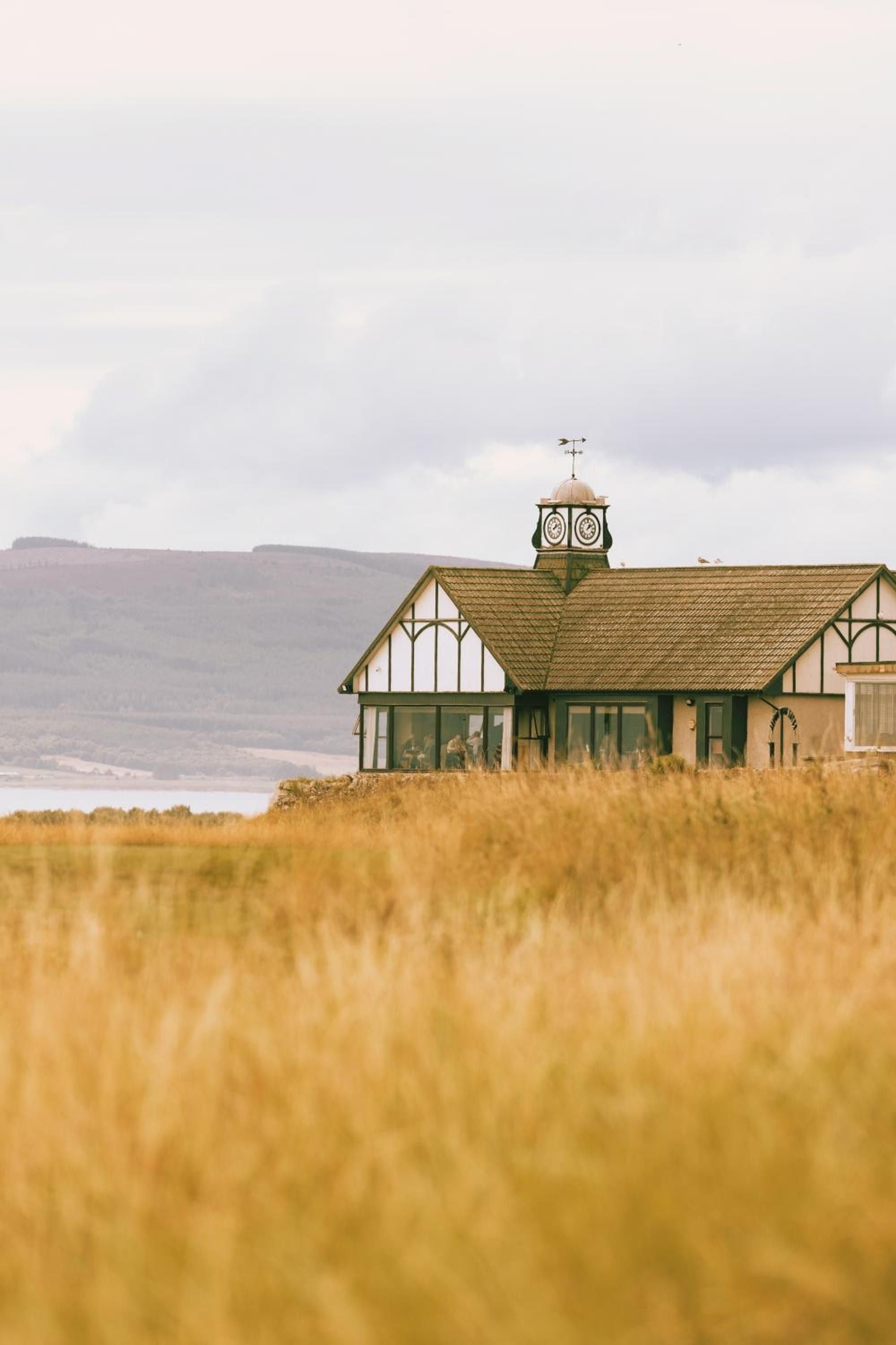 Tudor-style architecture emerges from golden coastal grasses, its weathered clock tower standing sentinel against Scotland's moody Highland backdrop and distant waters.