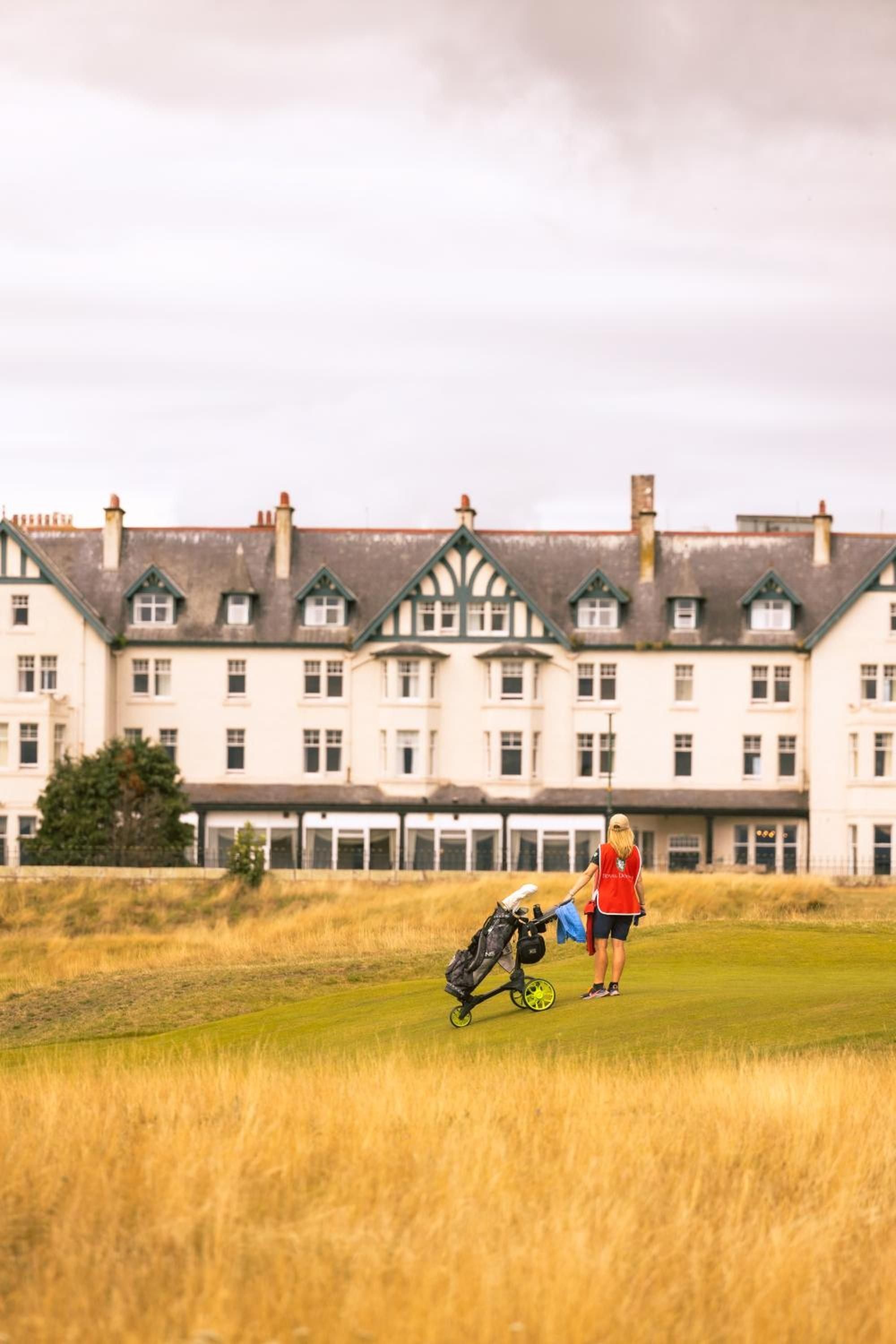 Golden afternoon light bathes the rough fairway as a golfer strolls past Dornoch Station's elegant Victorian façade, where Tudor-style gables frame the timeless Scottish highlands experience.