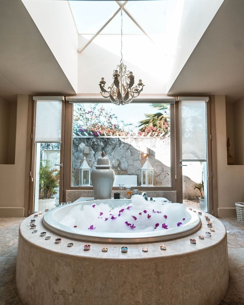 Purple orchid petals float in this sunken marble soaking tub, where candlelight dances beneath exposed beams and ocean breezes drift through panoramic windows.