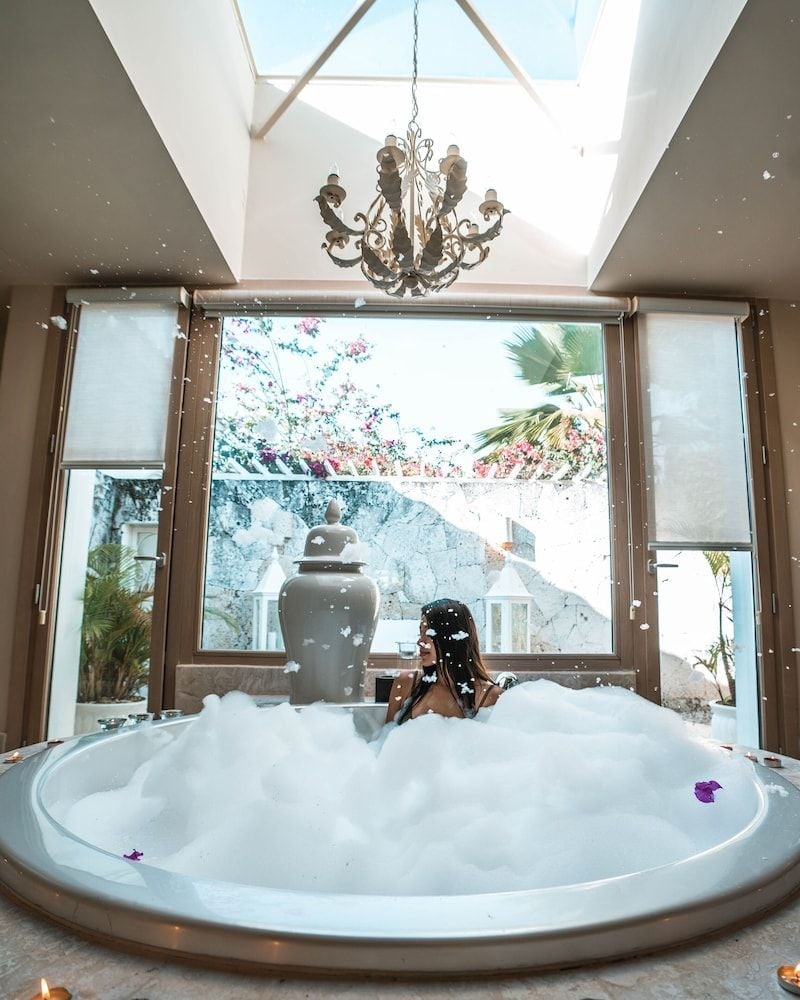 Rose petals drift like confetti through this opulent circular soaking tub, where Mediterranean light streams through floor-to-ceiling windows onto marble surfaces.