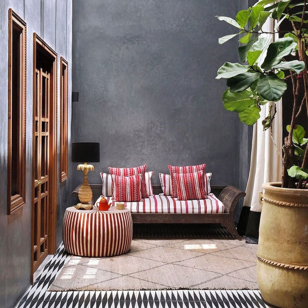 Moorish-inspired seating nooks showcase vibrant red-striped textiles against charcoal walls, while ornate wooden screens and terracotta planters create intimate moments of traditional Moroccan elegance.