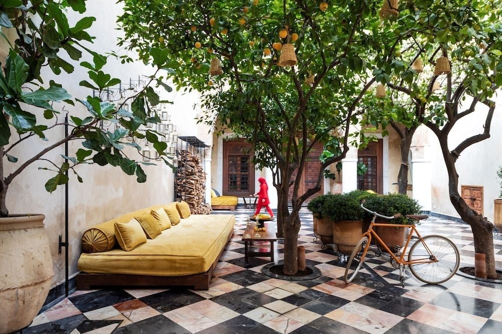 Citrus trees cast dappled shadows across checkered marble floors in this serene courtyard, where golden velvet cushions and vintage bicycles create an effortlessly chic Moroccan refuge.