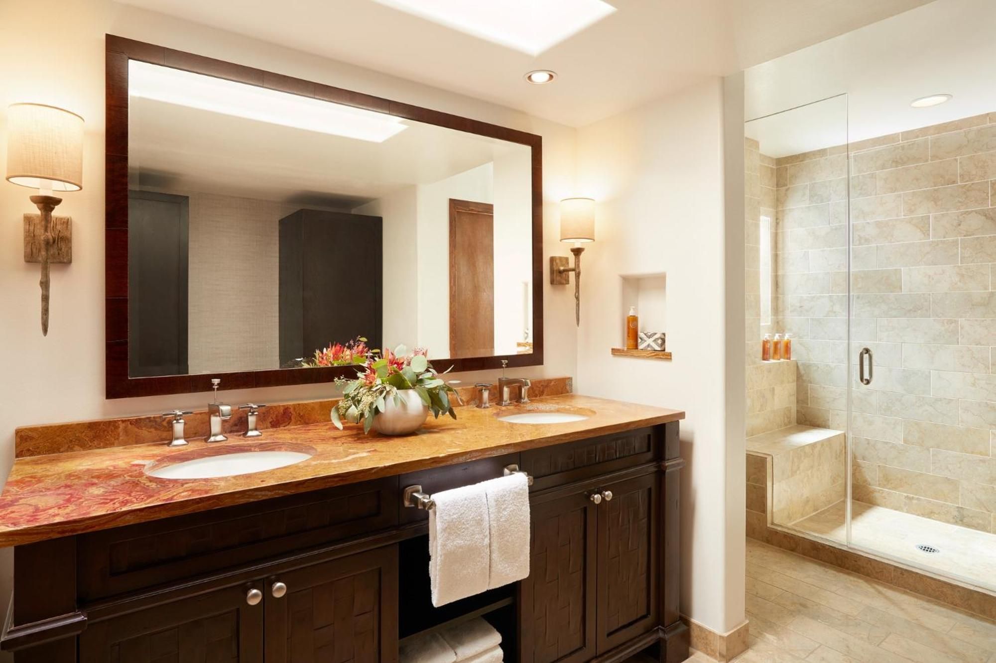 Desert-toned travertine and rich wood cabinetry create a spa-like sanctuary, where dual vanities and a glass-enclosed shower invite unhurried morning rituals.