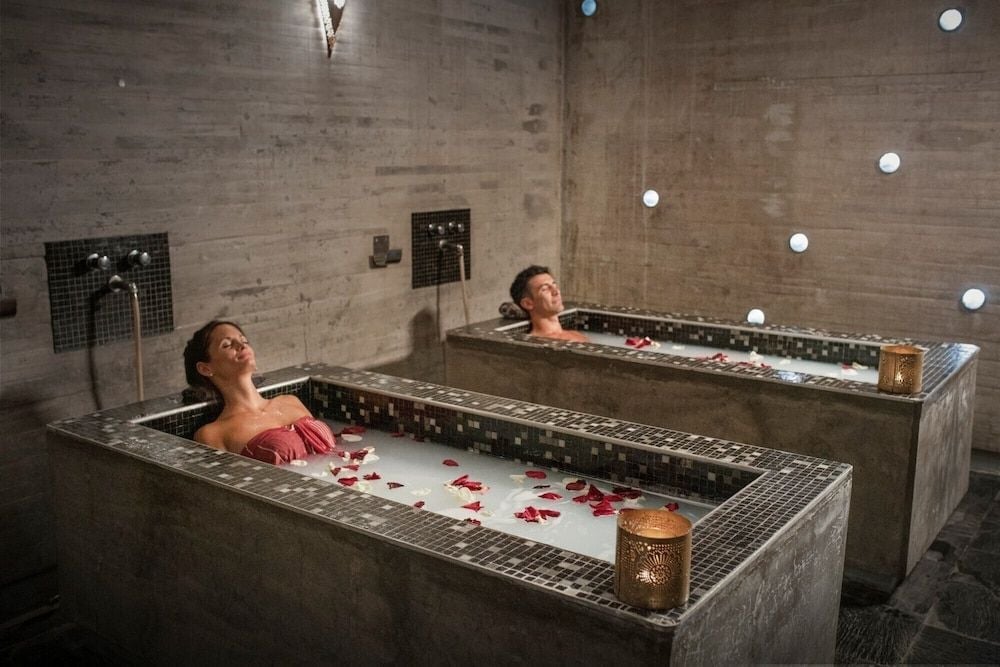 Couples unwind in side-by-side soaking tubs adorned with rose petals, while candlelight flickers against raw concrete walls in this intimate spa sanctuary.