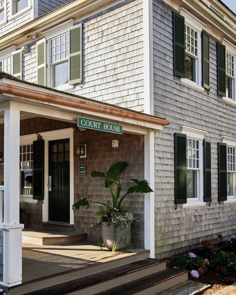 Weathered cedar shingles and forest-green shutters define this charming courthouse entrance, where dappled sunlight plays across the welcoming front porch and seasonal plantings.