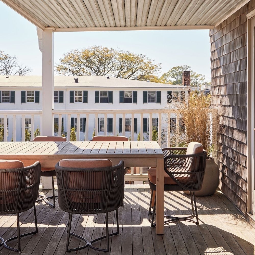 Weathered cedar shingles frame a sun-dappled terrace where wicker chairs gather around teak tables, overlooking classic Vineyard architecture through salt-kissed afternoon light.