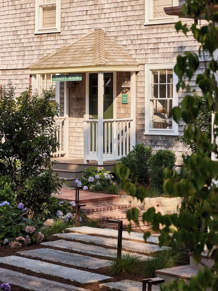 Weathered cedar shingles and hydrangea blooms frame the welcoming entrance to Chappy House, where brick pathways wind through manicured gardens toward classic New England charm.