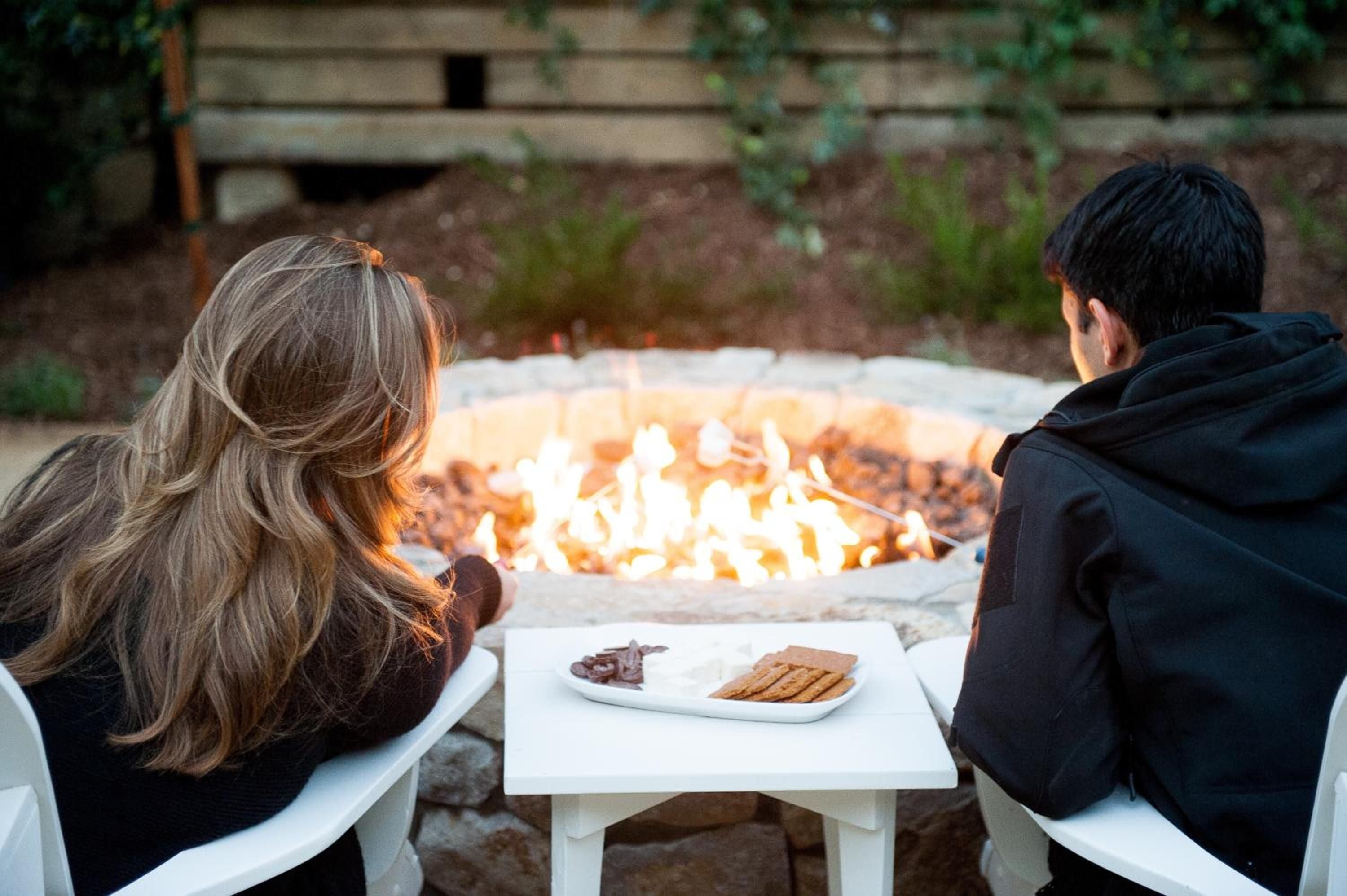 Couples gather around the crackling fire pit as evening light fades, sharing s'mores ingredients on crisp white tables amid Farmhouse Inn's intimate outdoor spaces.