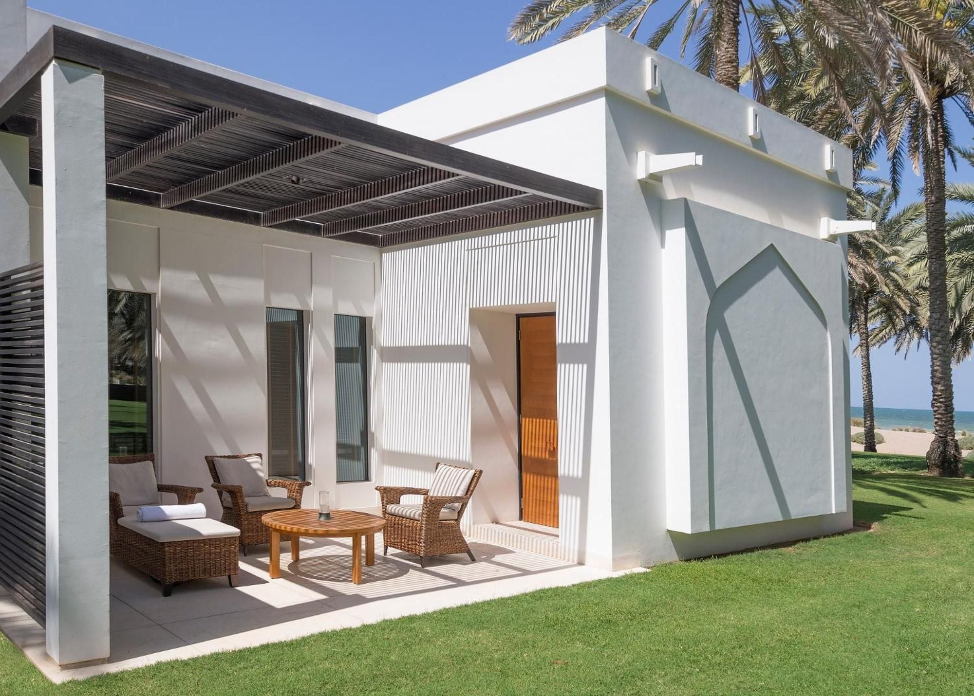 Wicker loungers nestle beneath slatted pergolas while palm shadows dance across manicured lawns, creating intimate outdoor sanctuaries steps from Muscat's pristine coastline.