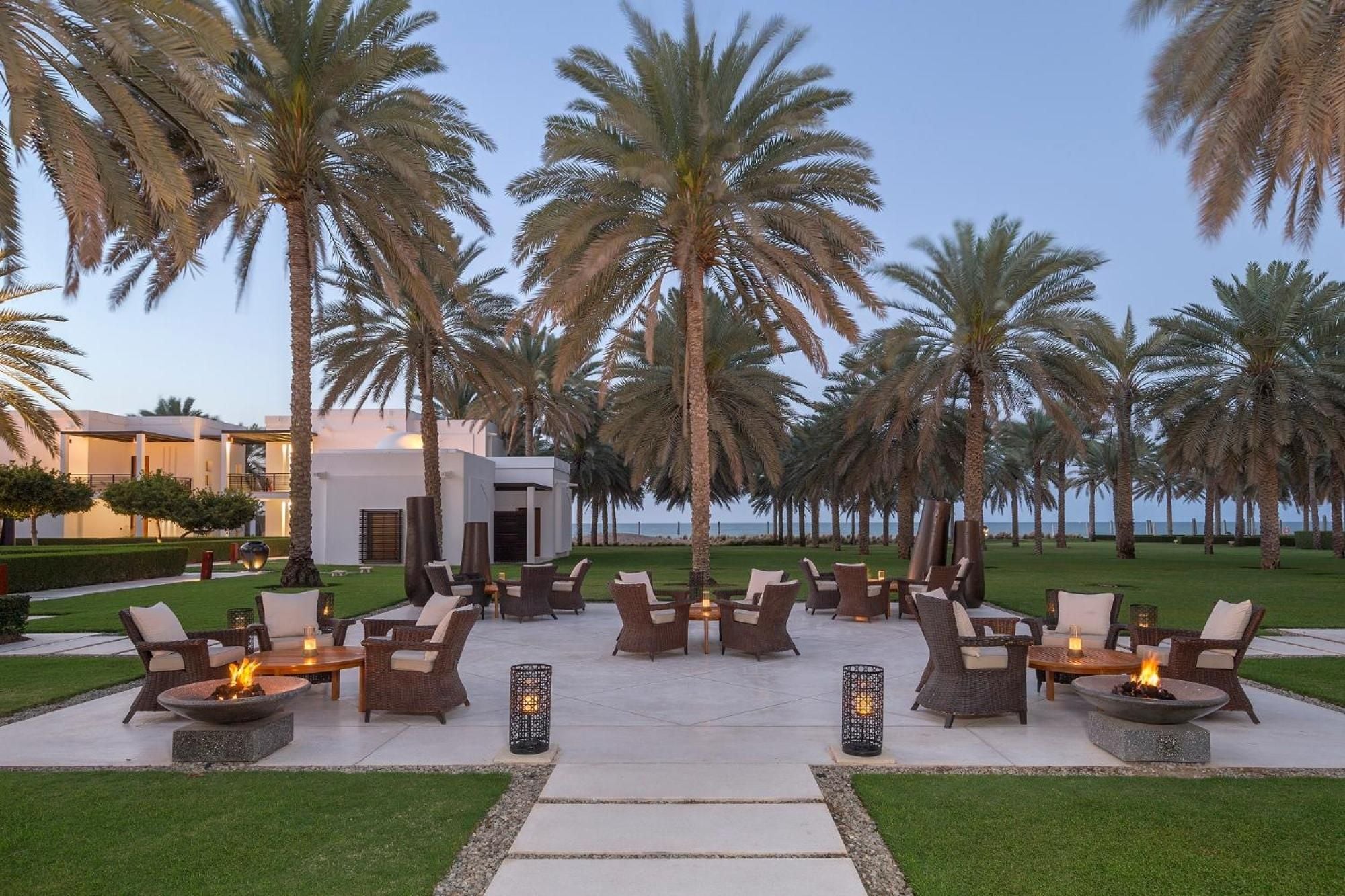 Flickering fire bowls and Moroccan-inspired lanterns illuminate wicker seating clusters beneath towering date palms, creating an enchanting desert oasis as twilight settles over manicured lawns.