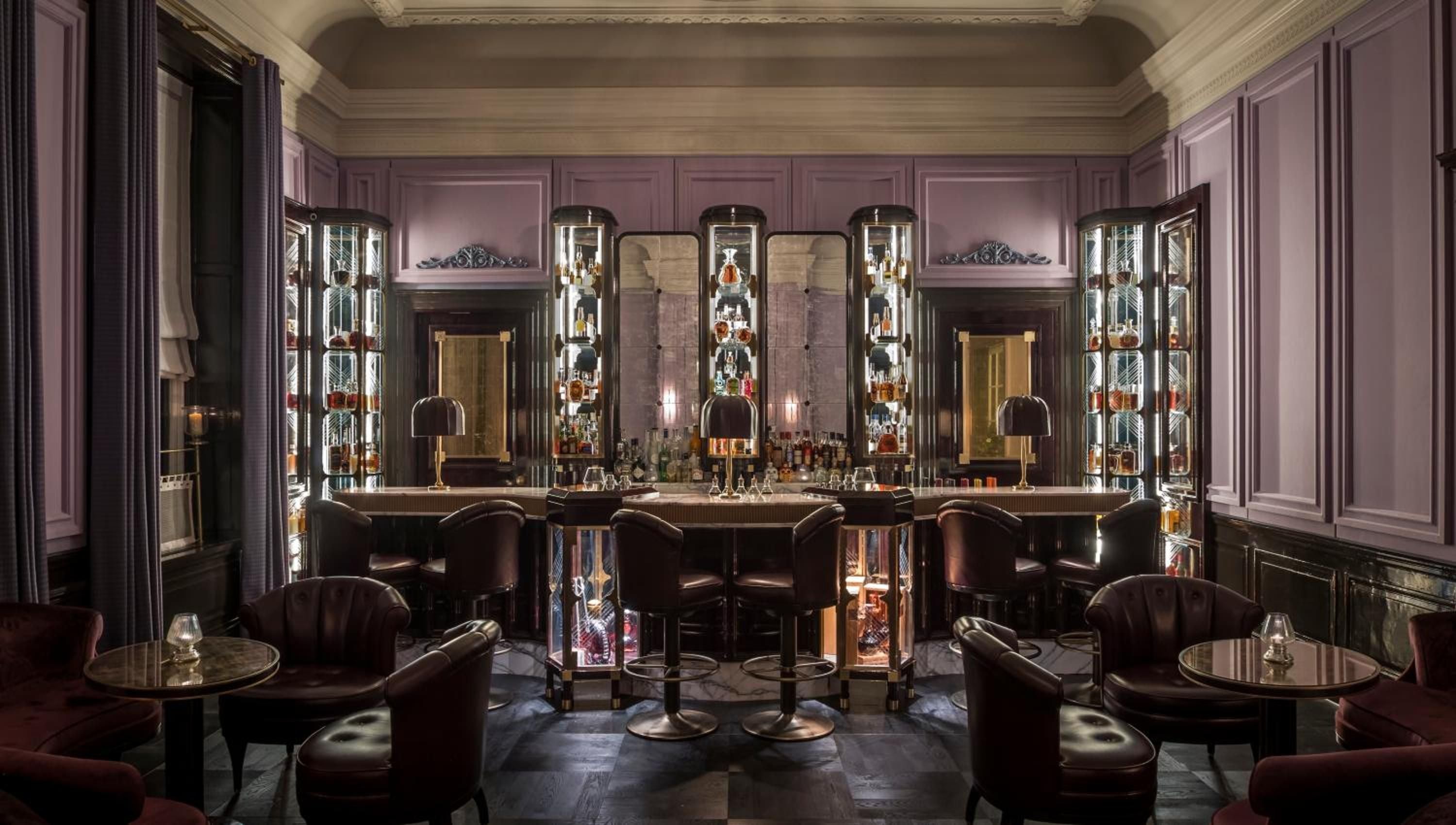 Purple-hued paneling and gleaming brass fixtures create an intimate speakeasy atmosphere, where vintage spirits line illuminated cabinets behind plush leather bar stools.
