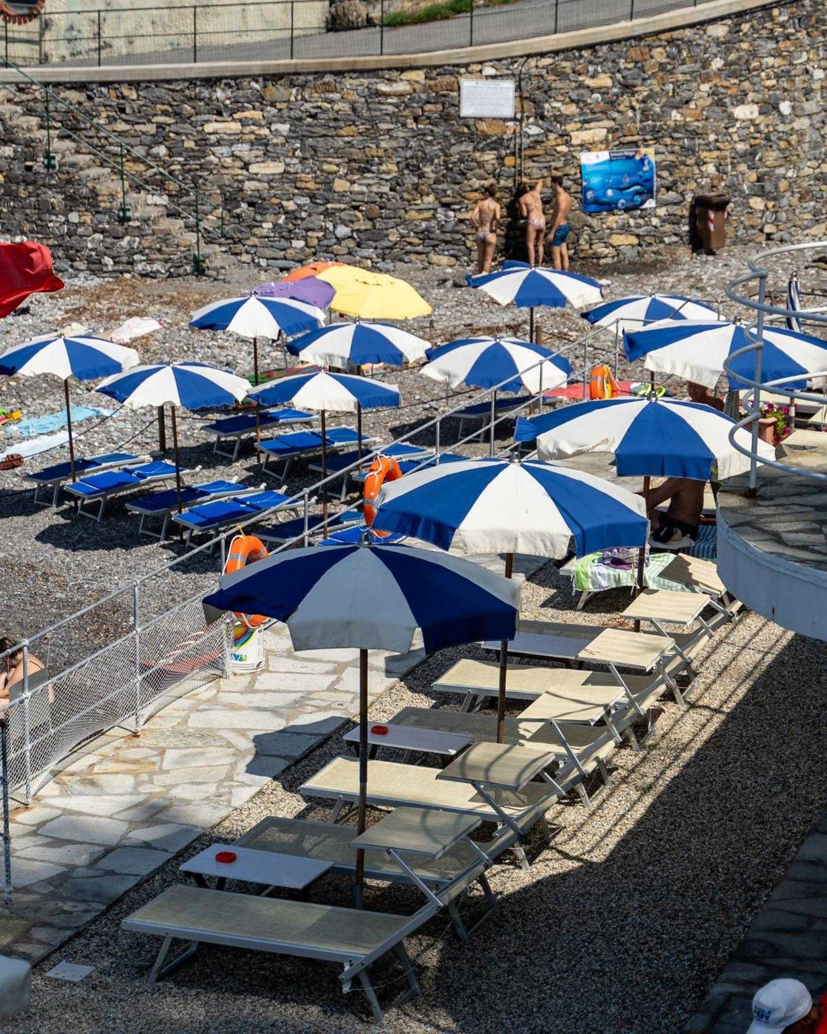 Blue and white striped umbrellas dot the pebbled shoreline, where ancient stone walls meet the Adriatic's crystalline waters in perfect Mediterranean harmony.