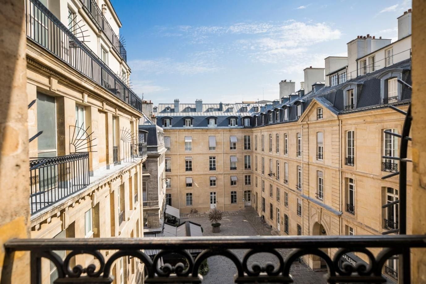 Honey-colored limestone facades frame a tranquil courtyard from your wrought-iron balcony, where Parisian rooftops stretch toward wispy afternoon clouds.