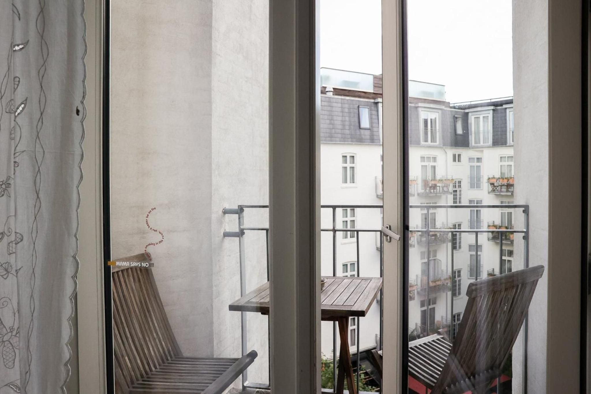 Wooden balcony chairs frame Copenhagen's historic rooftlines through floor-to-ceiling windows, where afternoon light filters across charming curtains and intimate outdoor seating.