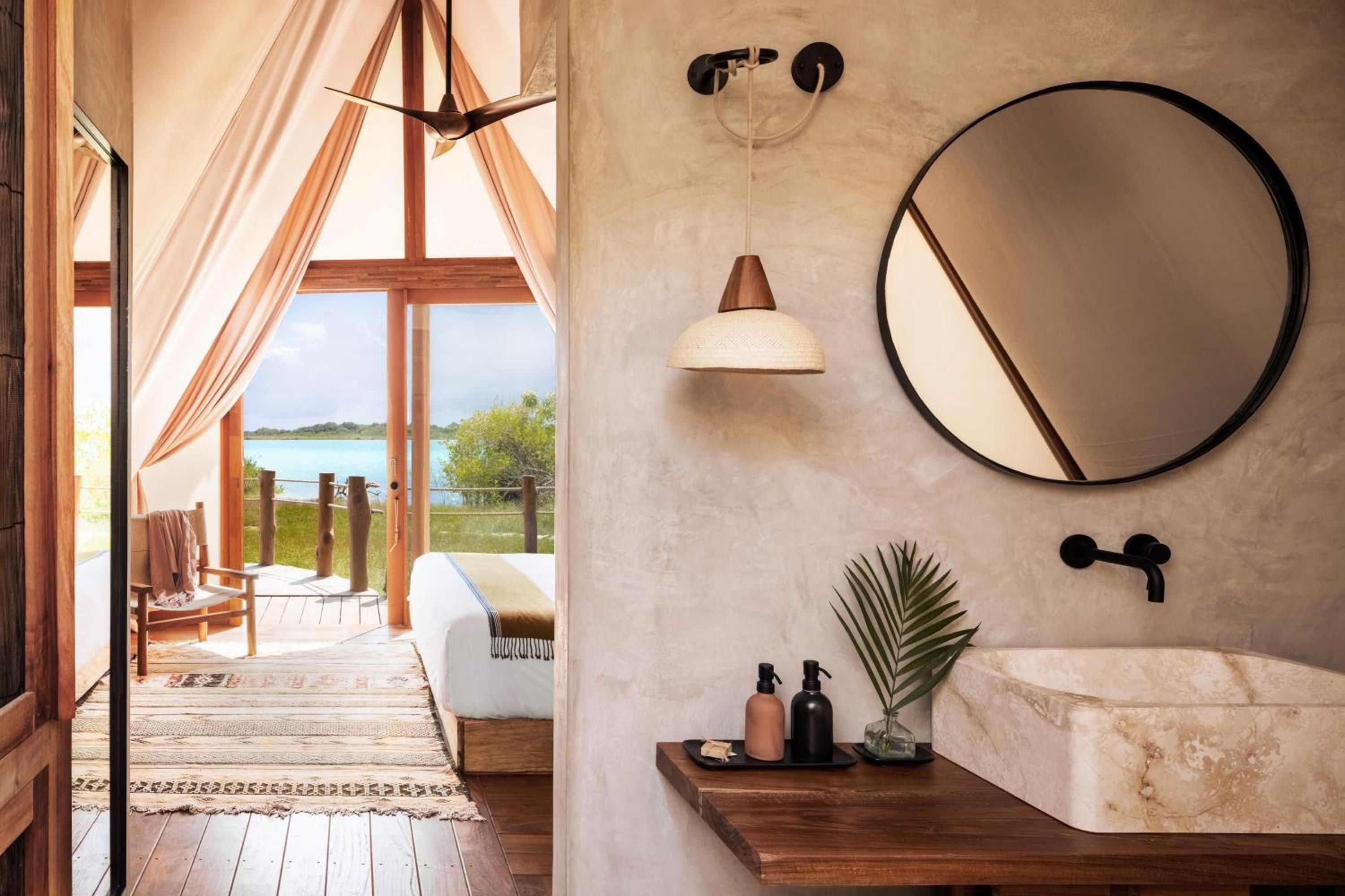 Natural limestone walls and warm wood complement black fixtures in this serene bathroom, where views of Bacalar's turquoise lagoon create an open-air sanctuary feeling.