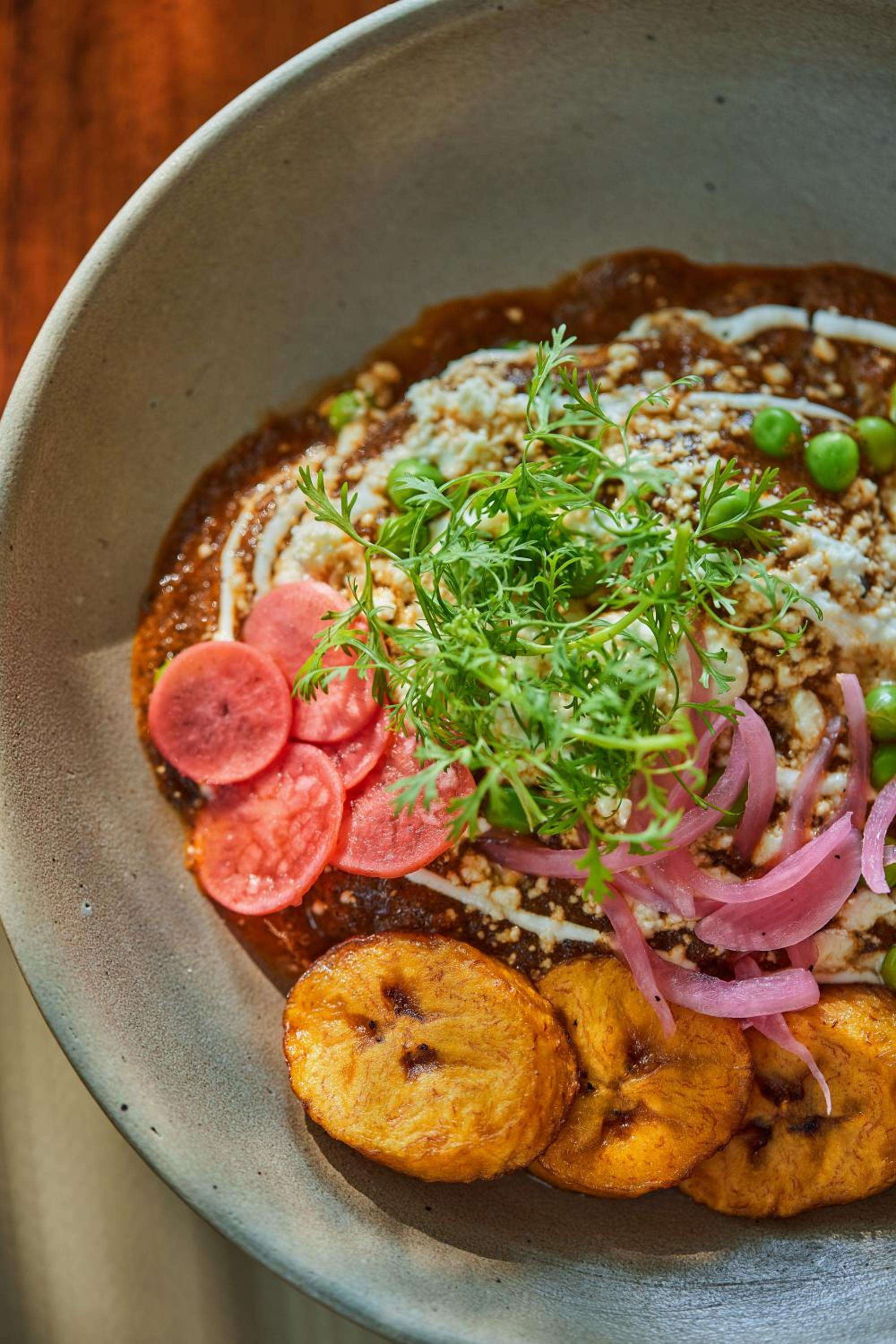 Artfully plated Mexican comfort food arrives in handcrafted ceramic bowls, with sliced radishes, fresh herbs, and golden plantains creating a feast for the senses.