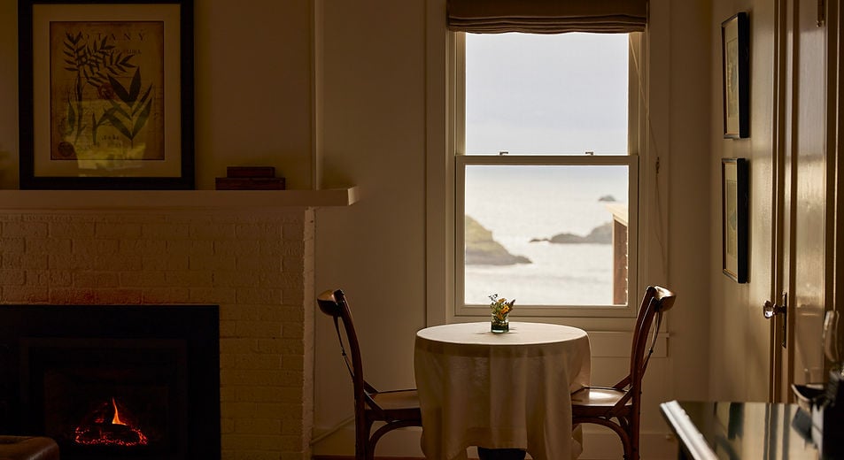 Golden afternoon light filters through oceanview windows, illuminating an intimate dining nook where glowing embers and coastal vistas create perfect sanctuary moments.