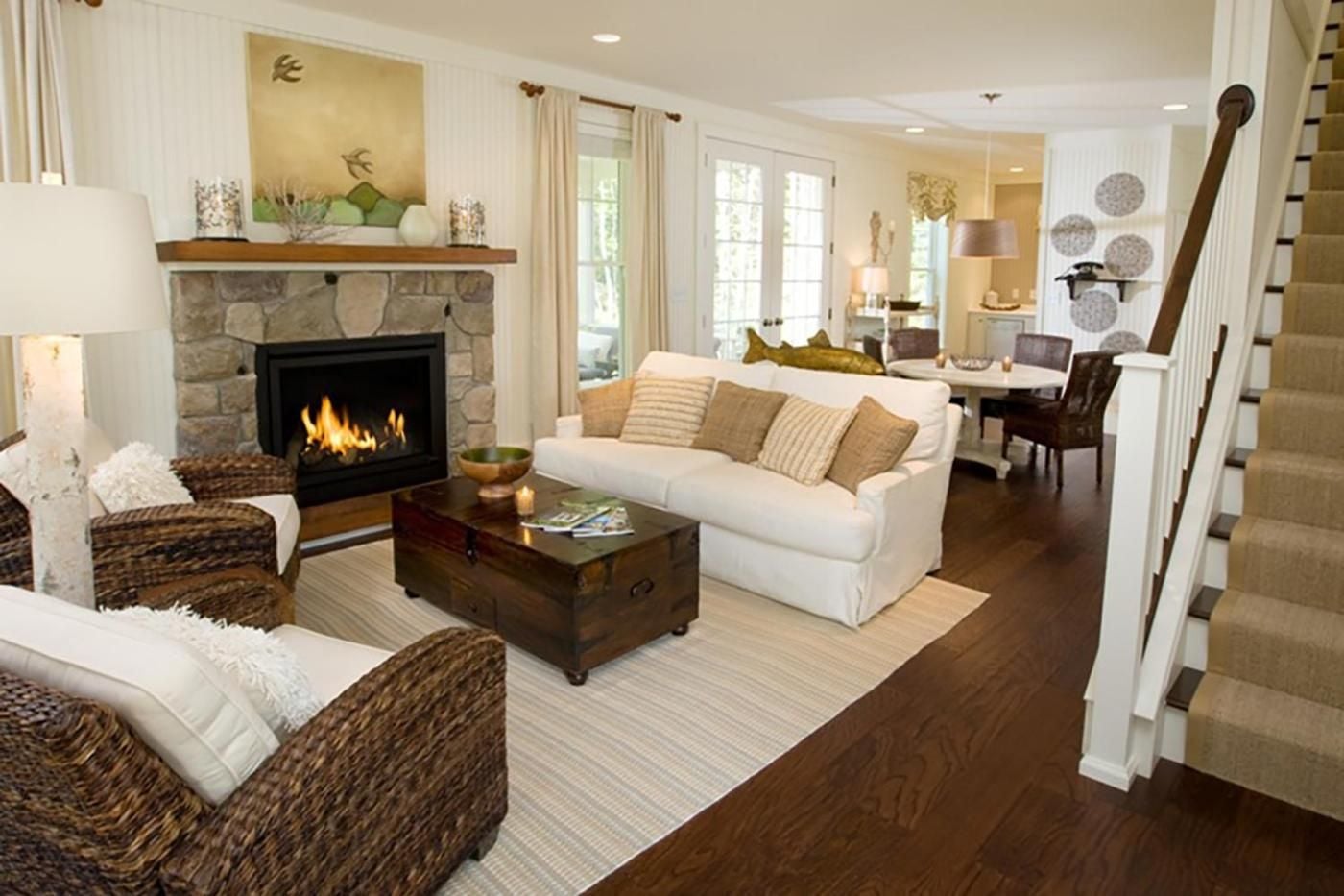 Wicker chairs and crisp white linens create coastal charm around the stone fireplace, where natural light streams through French doors onto gleaming hardwood floors.