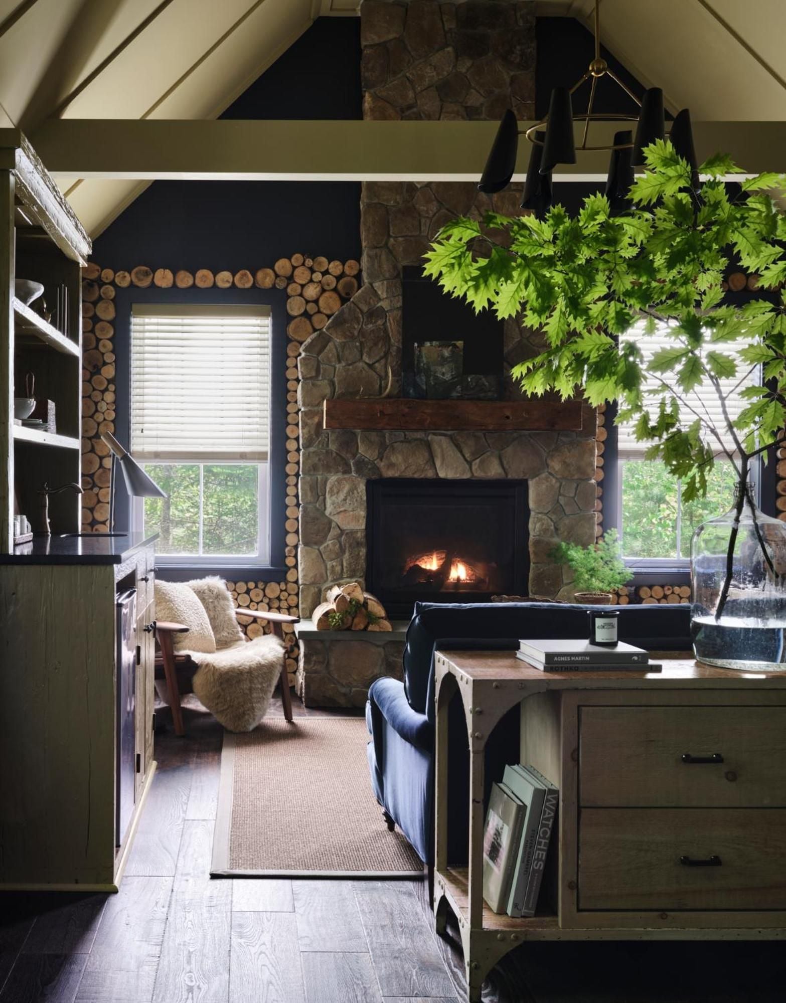 Stacked cordwood frames the stone fireplace in this cozy retreat, where exposed beams and a thriving potted tree create an inviting blend of rustic charm and modern comfort.