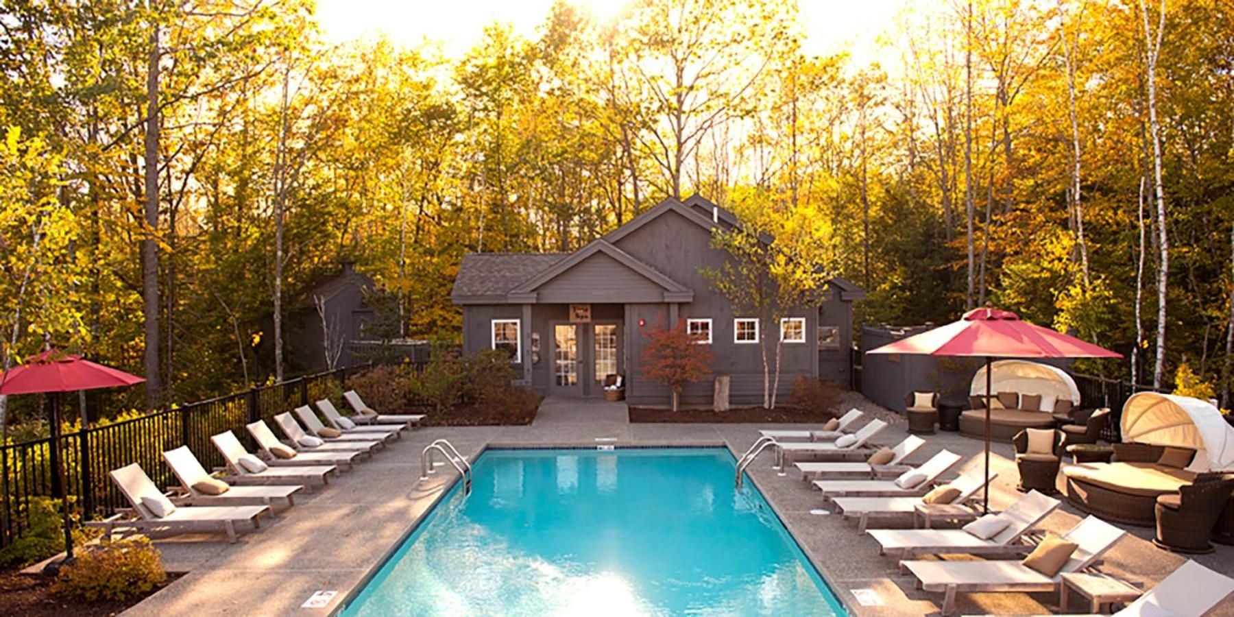 Autumn light filters through golden maples onto the heated pool, where weathered teak loungers and crimson umbrellas create an inviting refuge amid Maine's forest canopy.