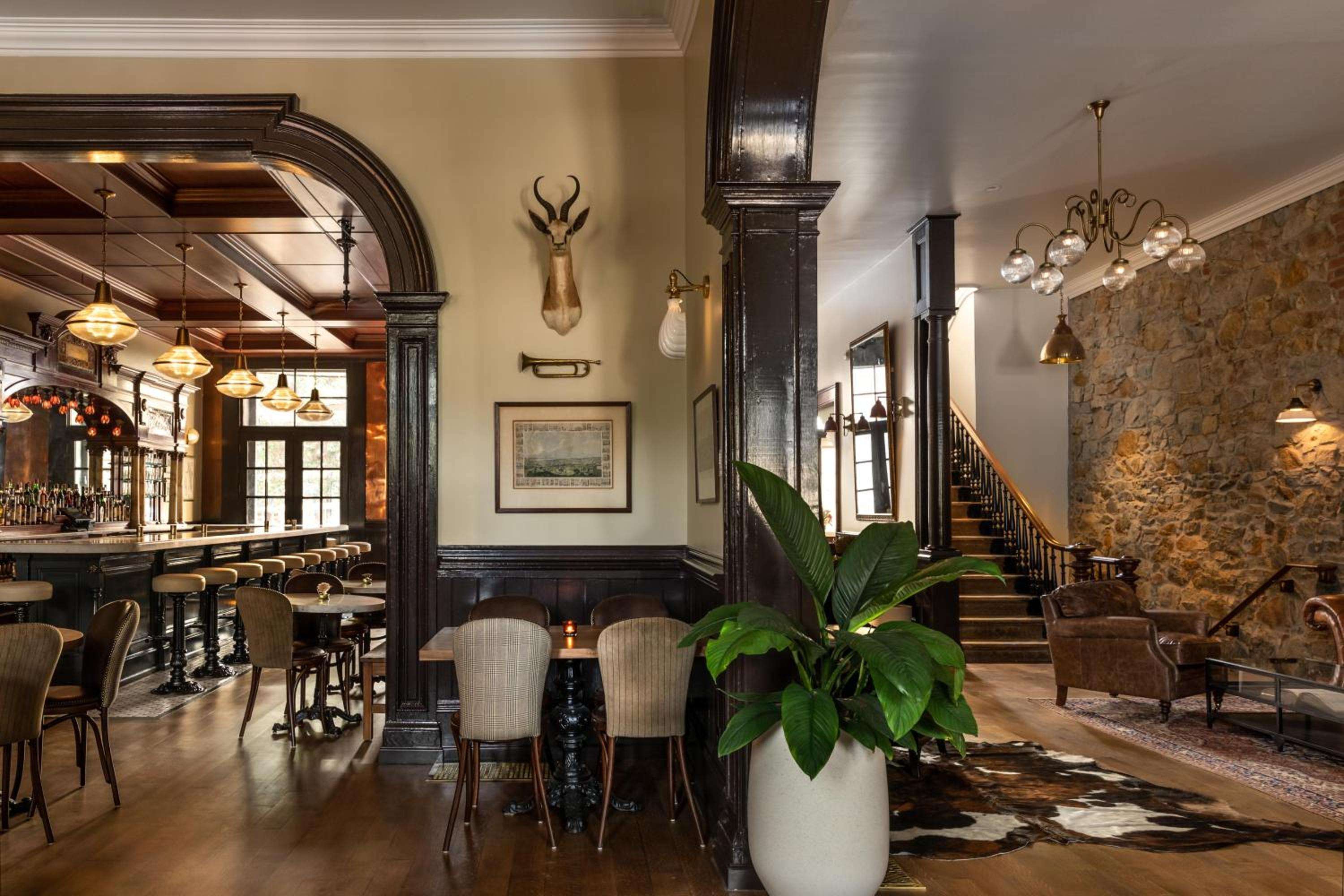 Dark wood columns and exposed stone create intimate dining alcoves, while pendant lights cast a golden glow over vintage-inspired tables and botanical accents.