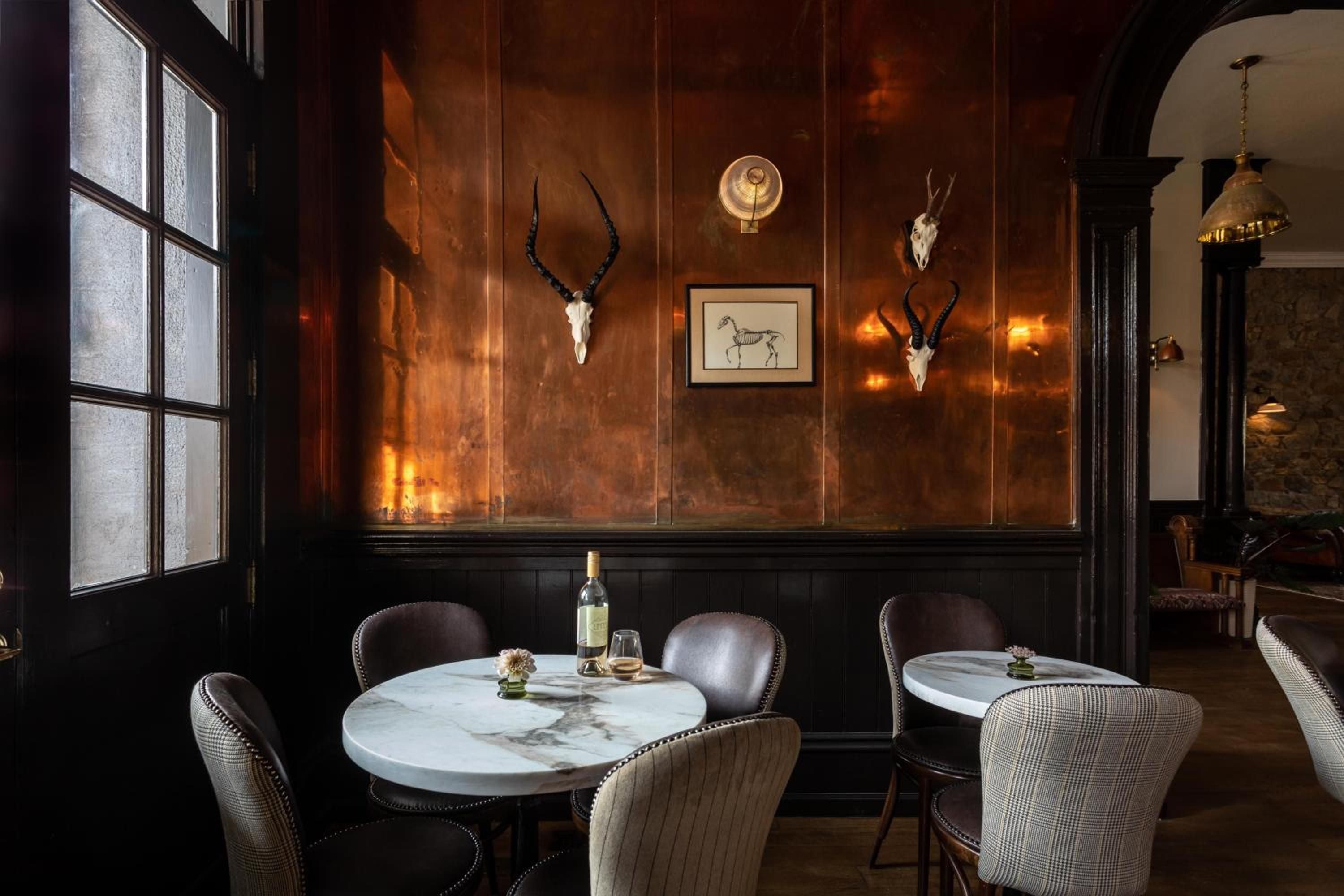 Copper-clad walls gleam beneath mounted antelope skulls in the intimate bar, where marble-topped tables and leather banquettes create an sophisticated hunting lodge atmosphere.