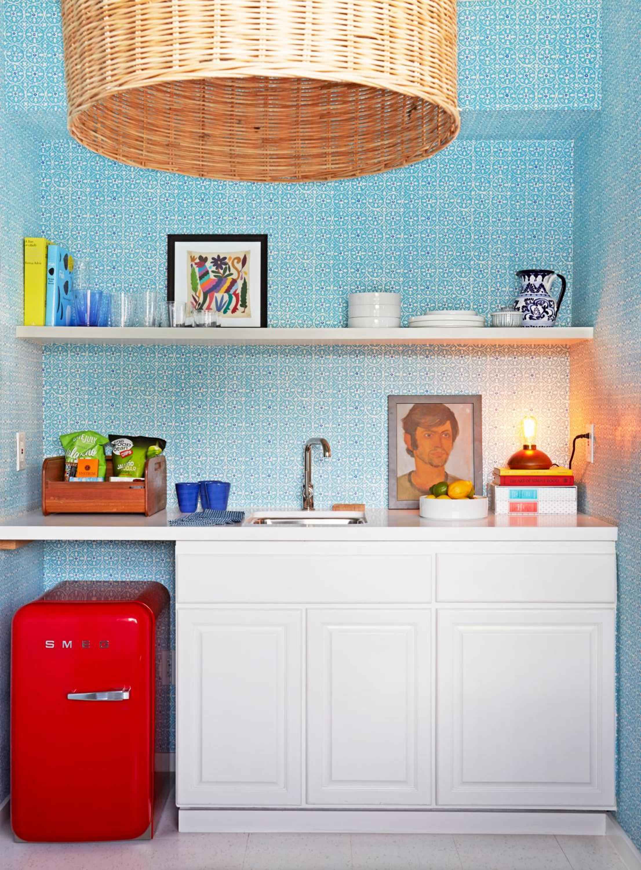 Whimsical blue geometric wallpaper and a cheerful red SMEG refrigerator create playful sophistication in this compact kitchenette, while wicker pendant lighting adds natural warmth overhead.