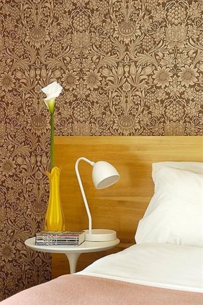 Intricate William Morris-inspired wallpaper creates an intimate backdrop for thoughtfully curated bedside vignettes, where white ceramic lamps cast gentle pools of reading light.
