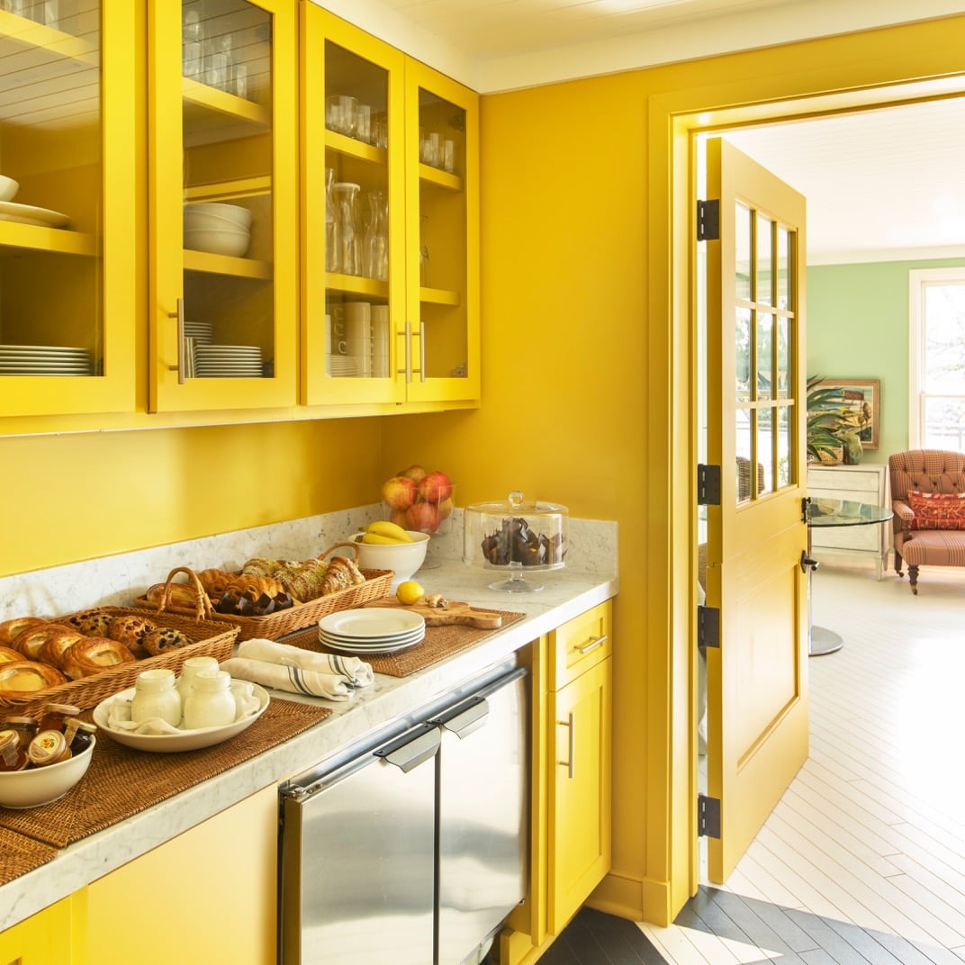 Sunny yellow cabinetry and marble countertops create a cheerful galley kitchen, where morning pastries and fresh fruit await in woven baskets beneath glass-front cupboards.