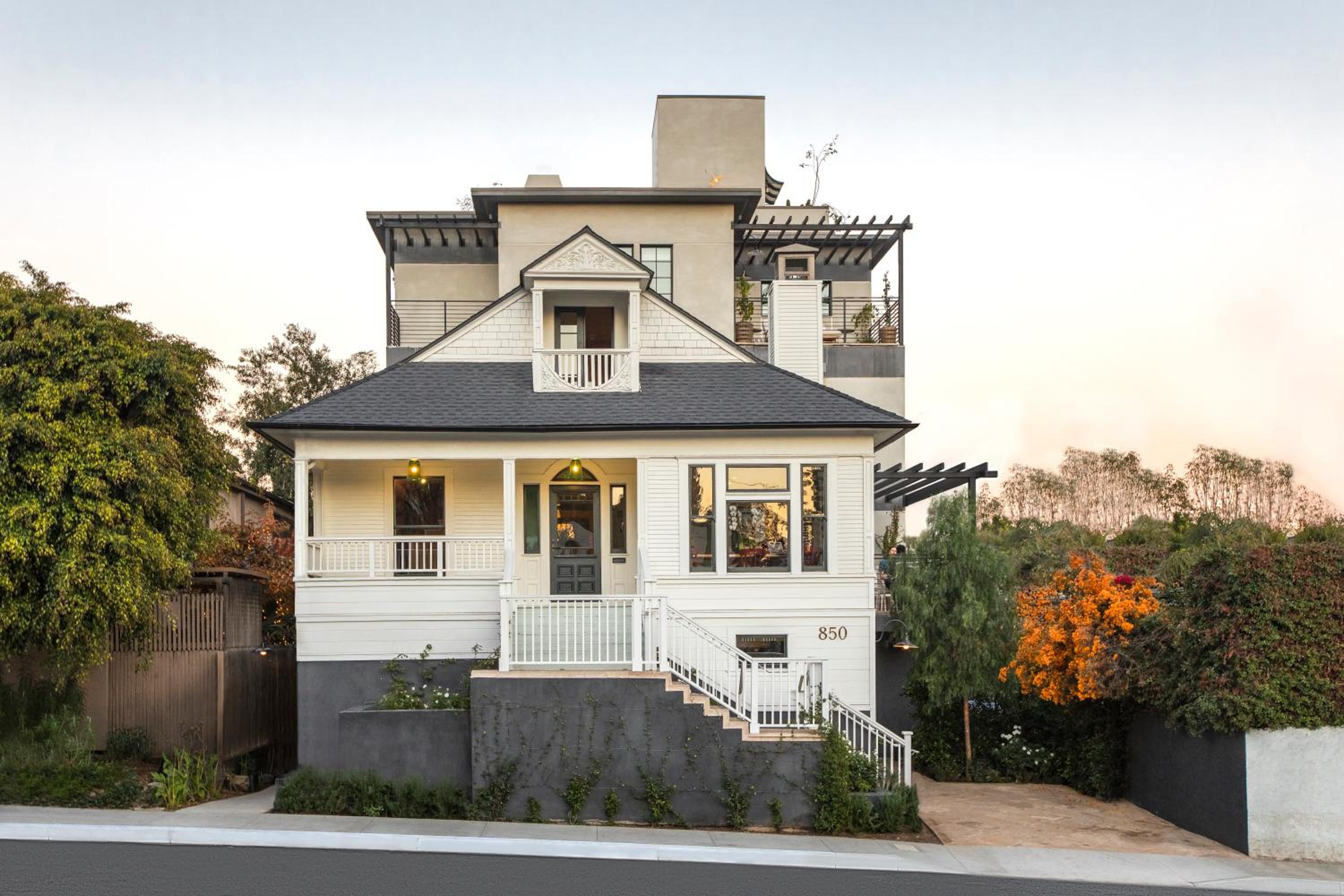 White clapboard charm meets modern luxury as this intimate hillside retreat rises above terraced gardens, its wraparound porch glowing warmly against the dusky sky.