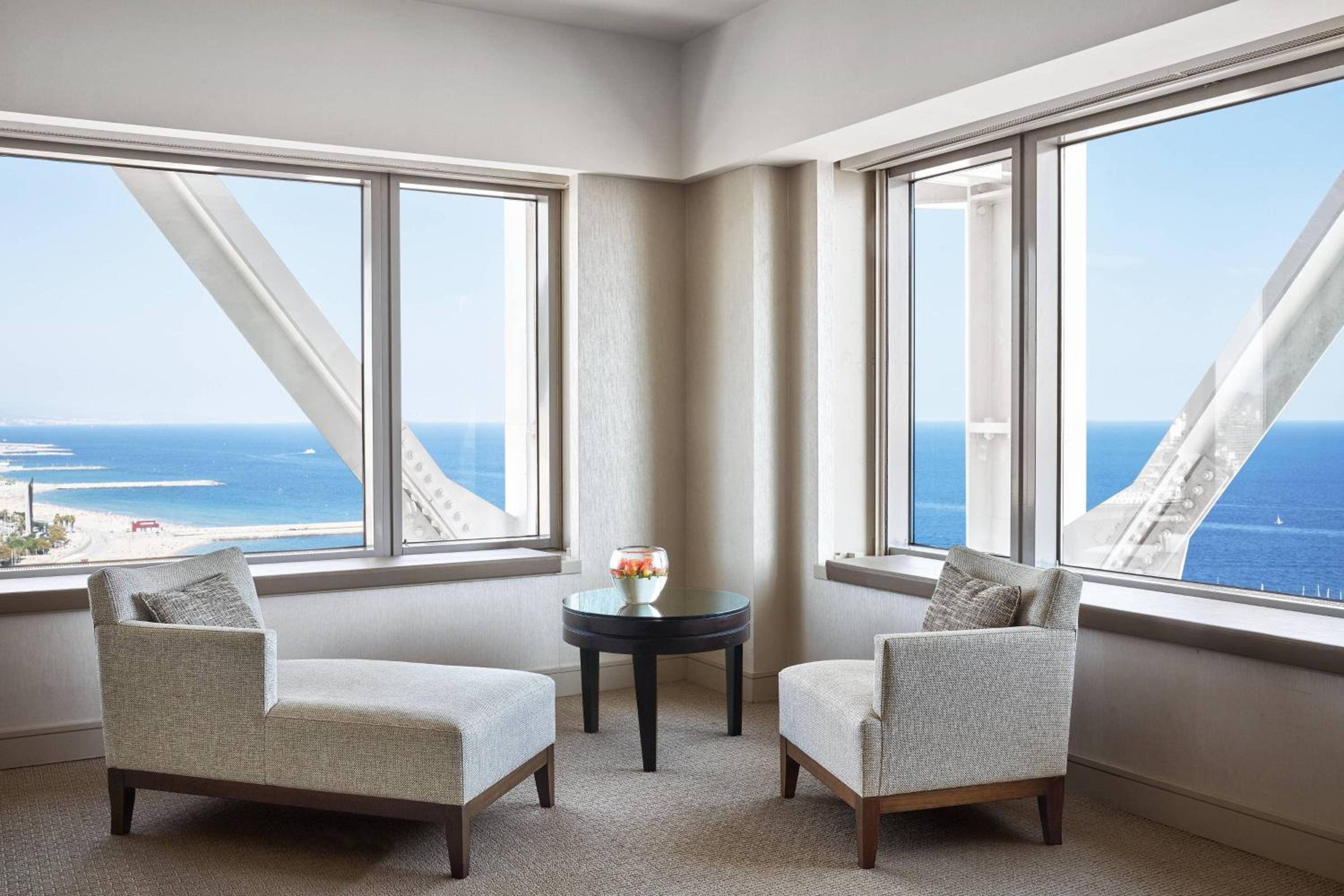 Corner windows frame the Mediterranean's endless blue while sculptural concrete beams create dramatic shadows across this serene seating area's neutral palette.