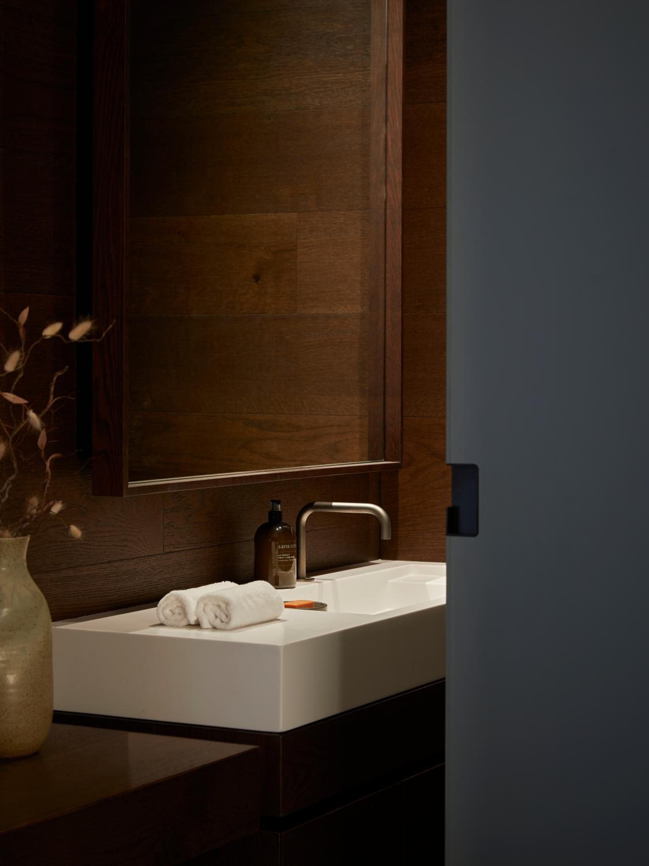 Warm timber frames the sleek basin where rolled towels and amber glass bottles create an intimate spa-like ritual in this thoughtfully appointed washroom.