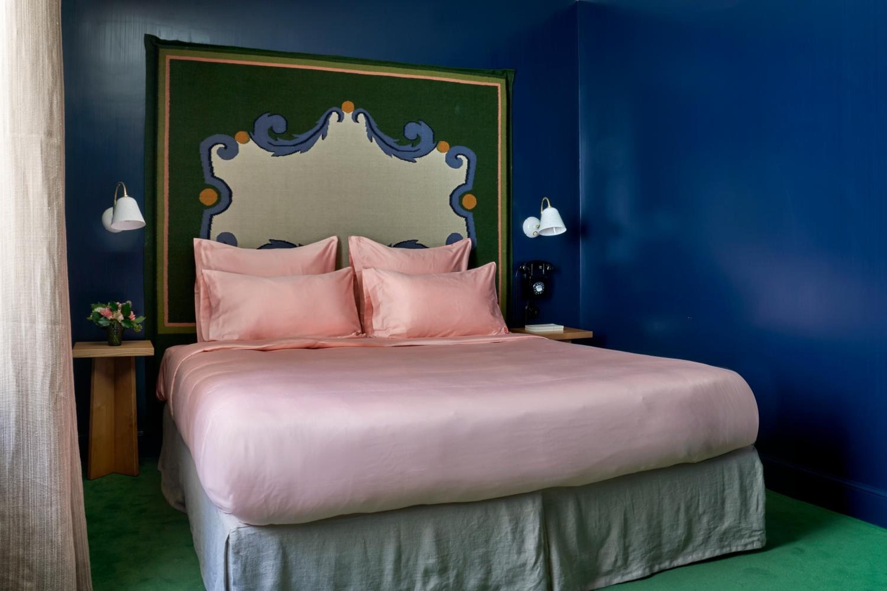 Coral-pink linens soften the dramatic navy walls, while an ornate painted headboard adds artistic flair to this intimate guest room bathed in warm sconce light.
