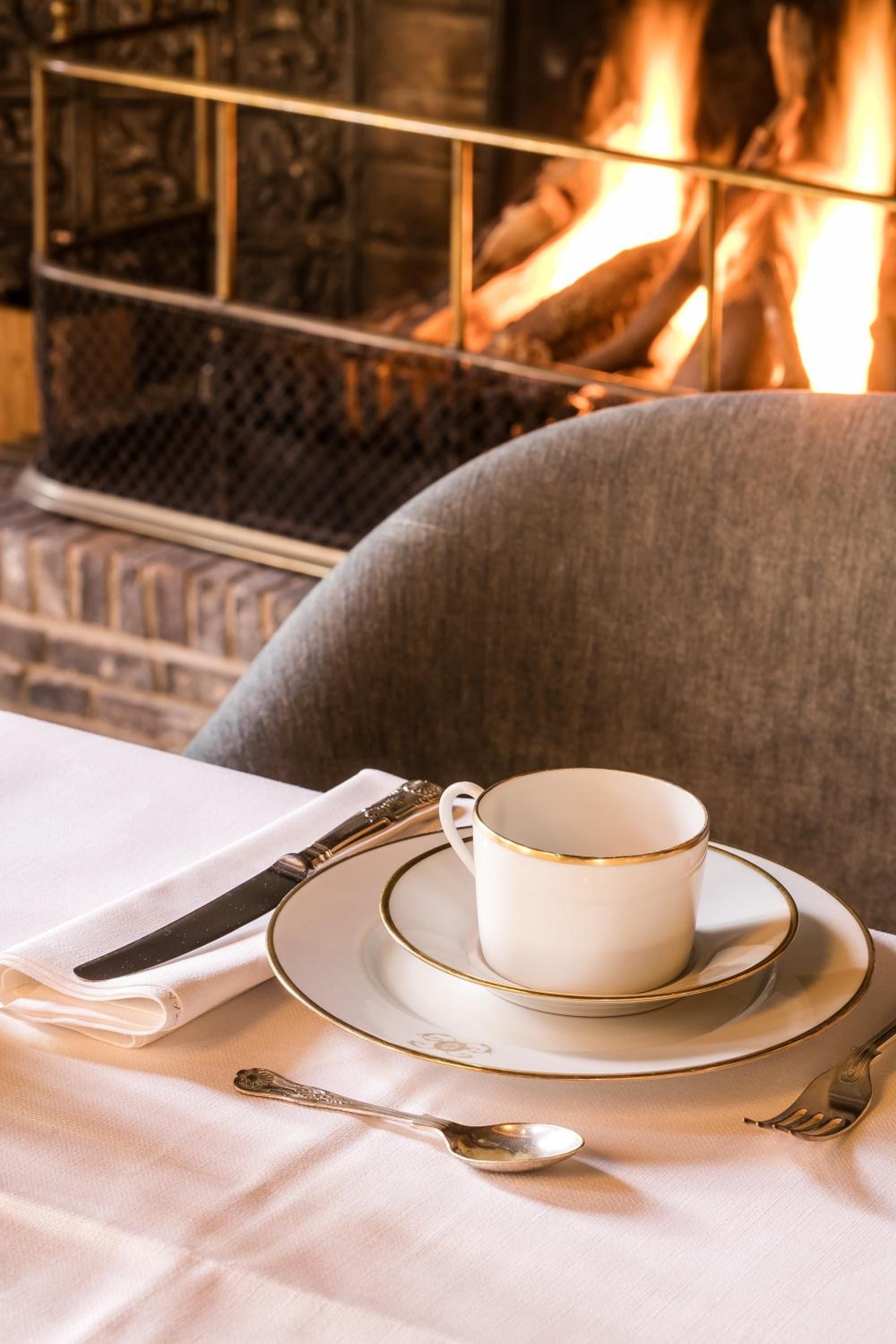 Elegant white porcelain with gold trim catches firelight at an intimate table, where ornate silverware and soft linens create an atmosphere of refined warmth.