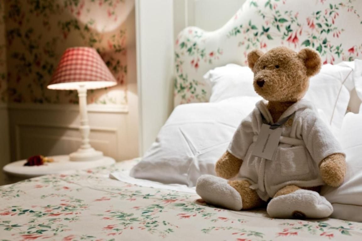 A beloved teddy bear in white pajamas sits sentinel beside floral wallpaper and gingham lampshades, embodying the hotel's endearing attention to whimsical comfort.