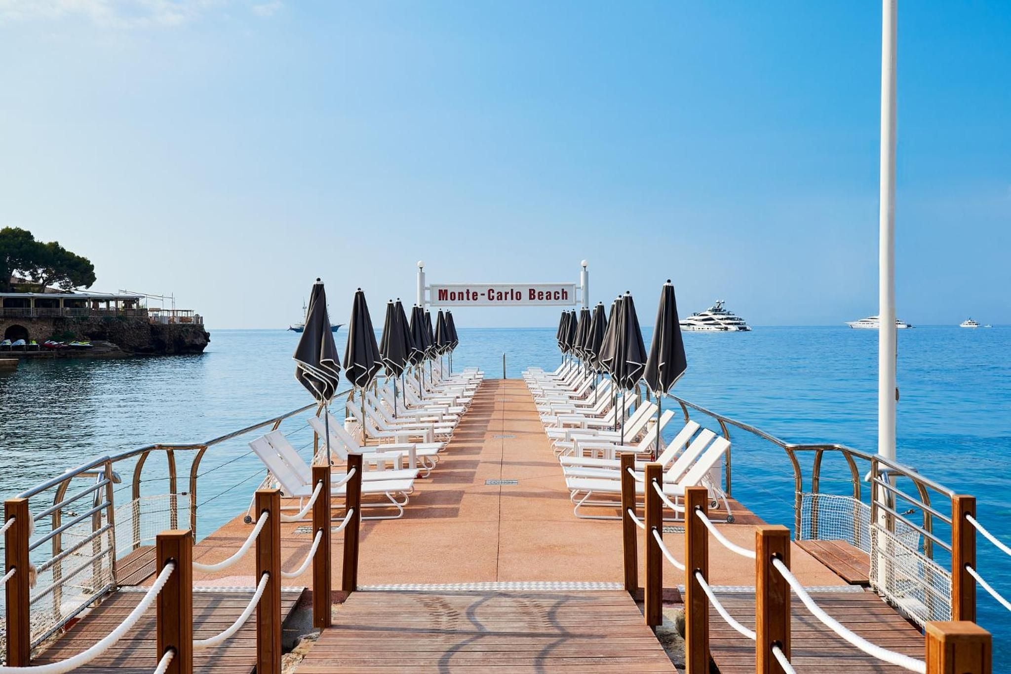 Monte-Carlo Beach's exclusive pier stretches into azure Mediterranean waters, lined with pristine loungers and furled umbrellas awaiting sun-seekers on the French Riviera.