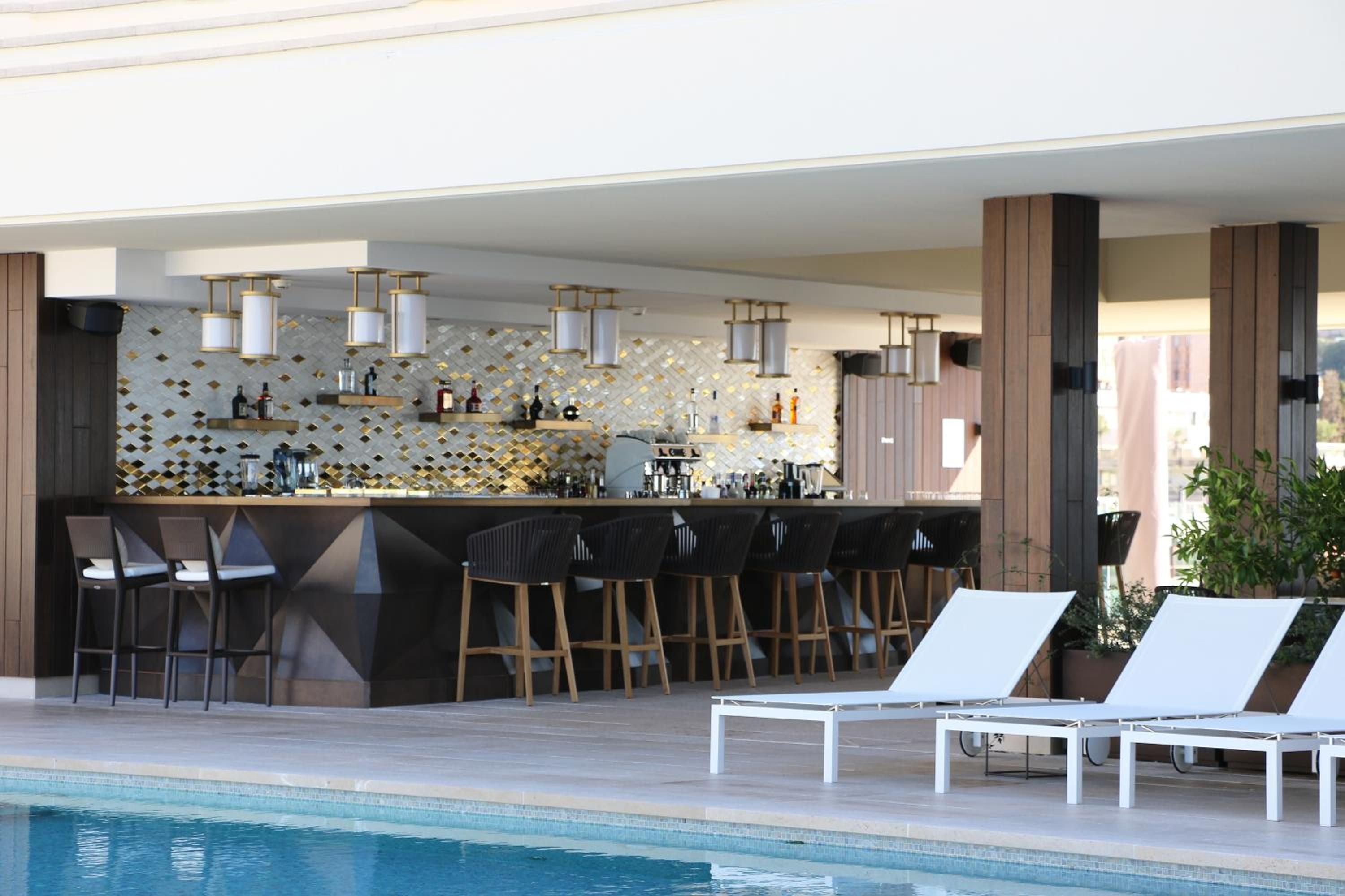 Geometric mosaic tiles gleam behind the poolside bar, where pendant lights cast warm shadows over sleek stools and crystal-clear water reflects the Riviera sun.