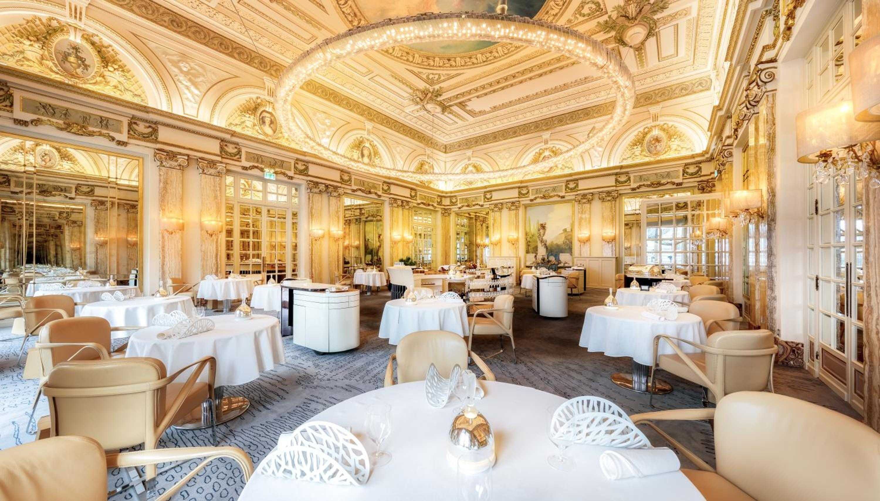 Gilded moldings and crystal chandeliers frame an opulent dining room where crisp white linens and contemporary chairs create an elegant juxtaposition of classic grandeur and modern comfort.