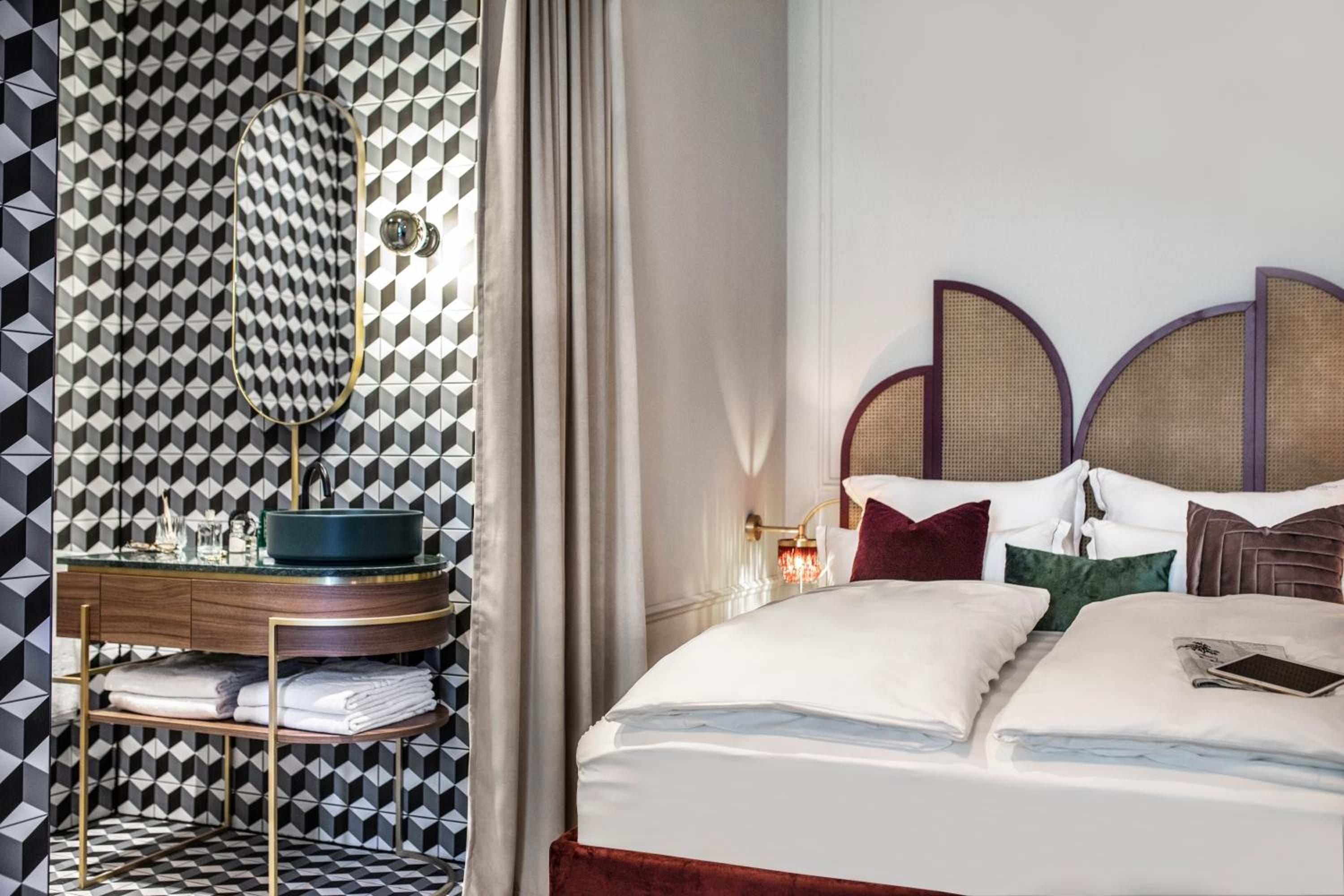 Geometric wallpaper creates dramatic contrast against cane-backed headboards and jewel-toned pillows, while brass fixtures and crisp linens complete this sophisticated retreat.