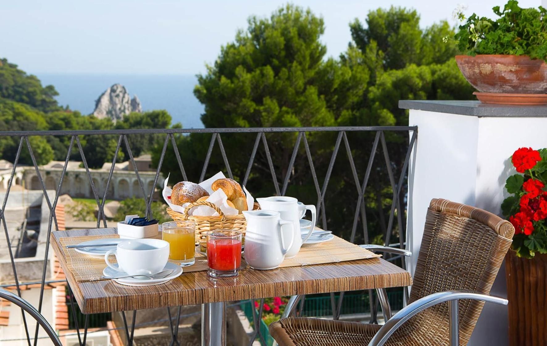 Morning sunlight illuminates a perfectly set breakfast table on the terrace, where warm pastries and fresh juice await against dramatic coastal cliffs and verdant Mediterranean foliage.