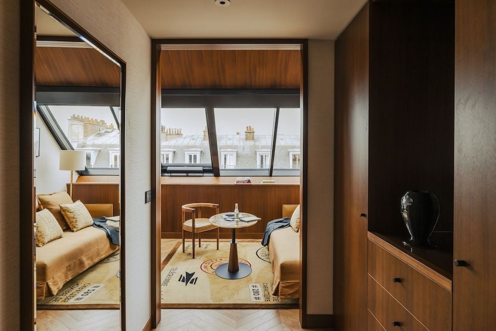 Warm walnut paneling frames Parisian rooftops through floor-to-ceiling windows, while honey-toned furnishings create an intimate living space that feels distinctly residential.