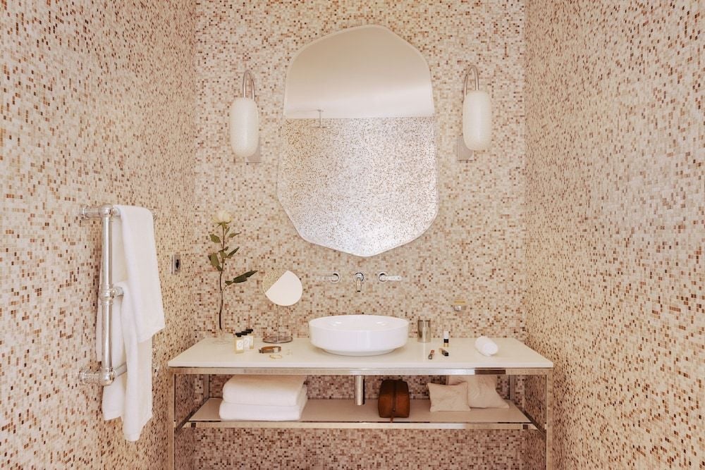 Honeyed mosaic tiles create a cocoon-like embrace around the sculptural vanity, where pendant lights cast a warm glow over vessel sink and organic mirror.